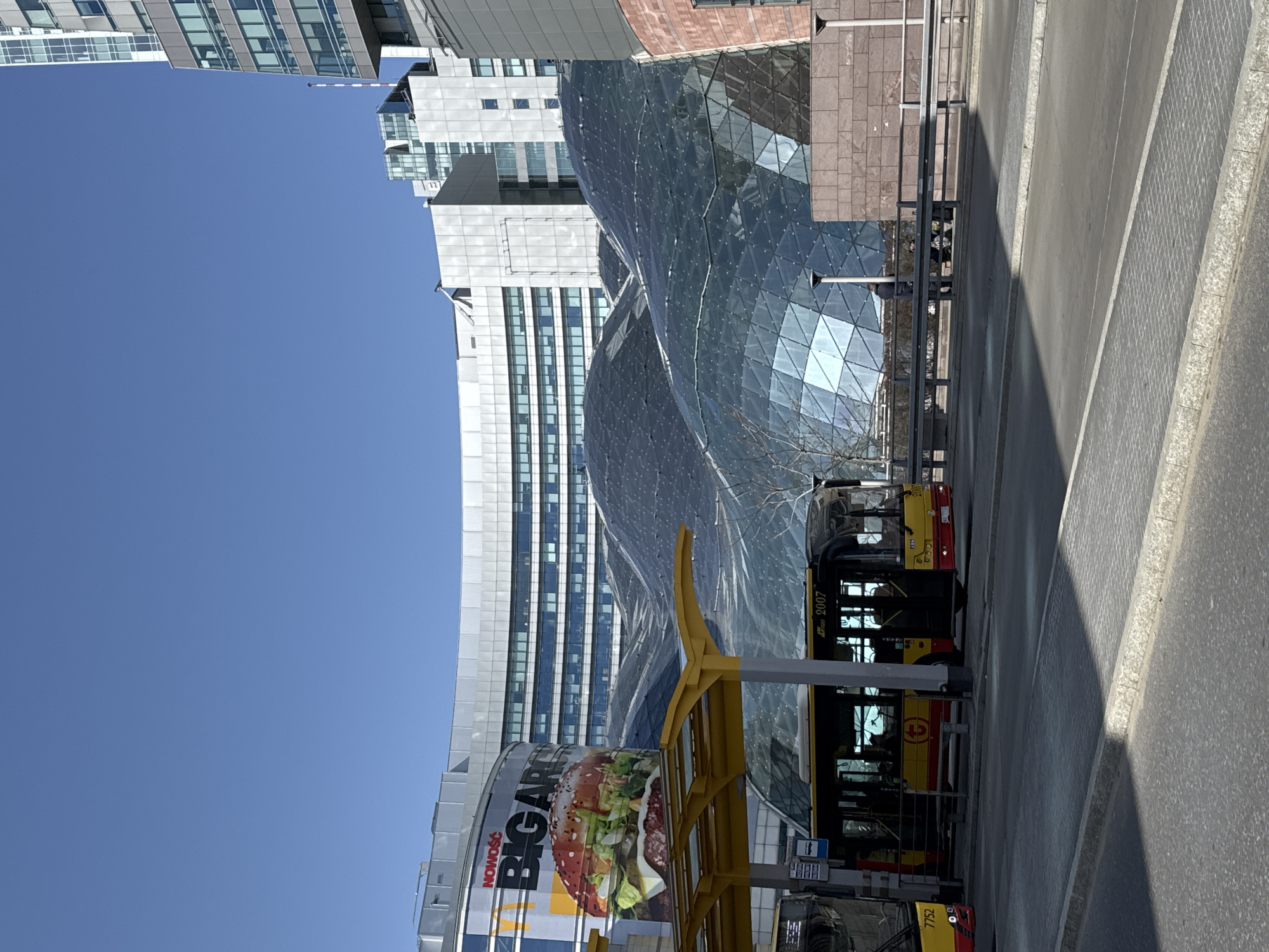 The image shows a street scene in Warsaw, Poland, dominated by modern architecture and public transportation. Visible are a bus stop with buses, one labeled 7752, featuring the Warsaw city public transport logo, a red circle with a white "1" in the center. A large advertisement for "BIG ART" is on a building, featuring a burger. Behind the bus stop, a modern glass structure with geometric facets is evident, suggesting a business or cultural center. Tall, white buildings and clear blue skies complete the urban landscape.