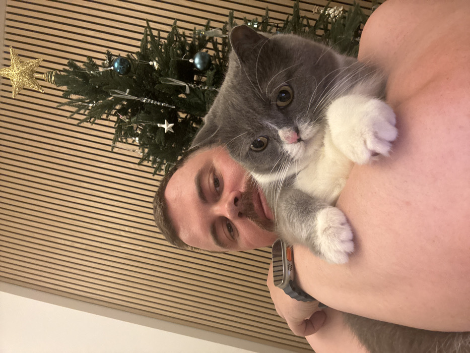 An adult male, approximately 30-40 years old with short brown hair and a mustache, is depicted in a close-up, chest-up shot. He has light skin and is bare-chested, wearing a dark-colored smartwatch with an orange accent on his left wrist. He holds a short-haired bicolor cat, predominantly grey with white fur on its chest, paws, and face, against his right shoulder and upper chest. The cat has amber eyes and a pink nose, with its front paws resting on the man's skin. Both the man and the cat are looking directly forward, toward the camera.
The background consists of a vertically slatted light-brown wooden wall, with a plain white wall section visible to the upper left. To the right, a small-to-medium-sized artificial Christmas tree is partially visible, adorned with a golden star topper, blue spherical ornaments, white star-shaped ornaments, and clear icicle-shaped ornaments. The scene is set indoors, within a residential or hotel environment in Dubai, United Arab Emirates.