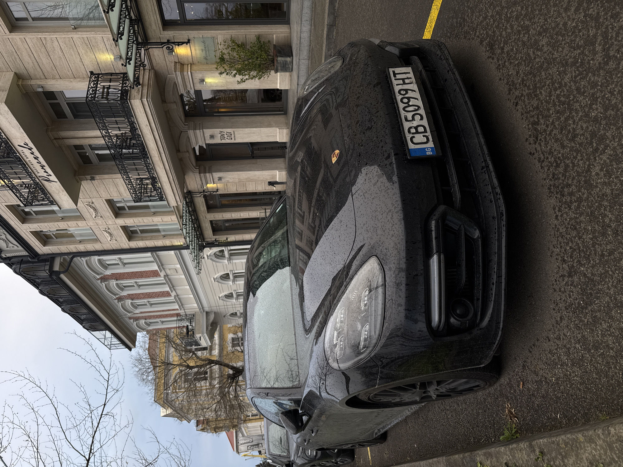 The image shows a black Porsche Panamera parked on a wet street in Burgas, Bulgaria. The car's license plate reads "CB 5099 HT." The backdrop features buildings with classical architectural details and wrought iron balconies. One building has a sign that reads "TONY GIGI." The weather appears overcast.