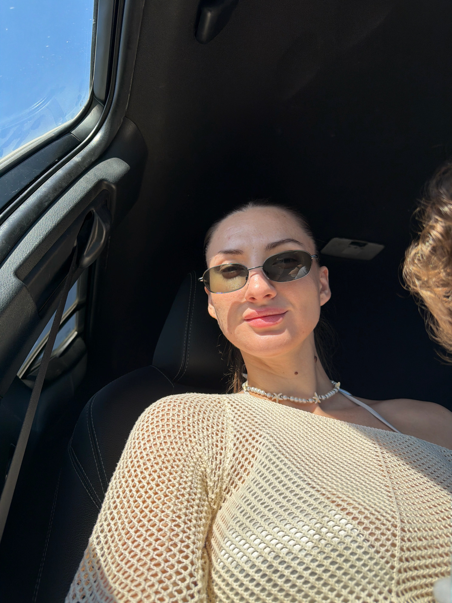 A woman, wearing sunglasses and a beige mesh top over a white bra, is seated in a car. She has a pearl necklace with starfish charms around her neck. The car interior, including a black seat and part of the dashboard, is visible. Sunlight streams in from the window, illuminating her face and outfit. The background is dark. This image does not provide any information or context to determine if it is located in Sharm El Sheikh, Egypt, or any other specific city.