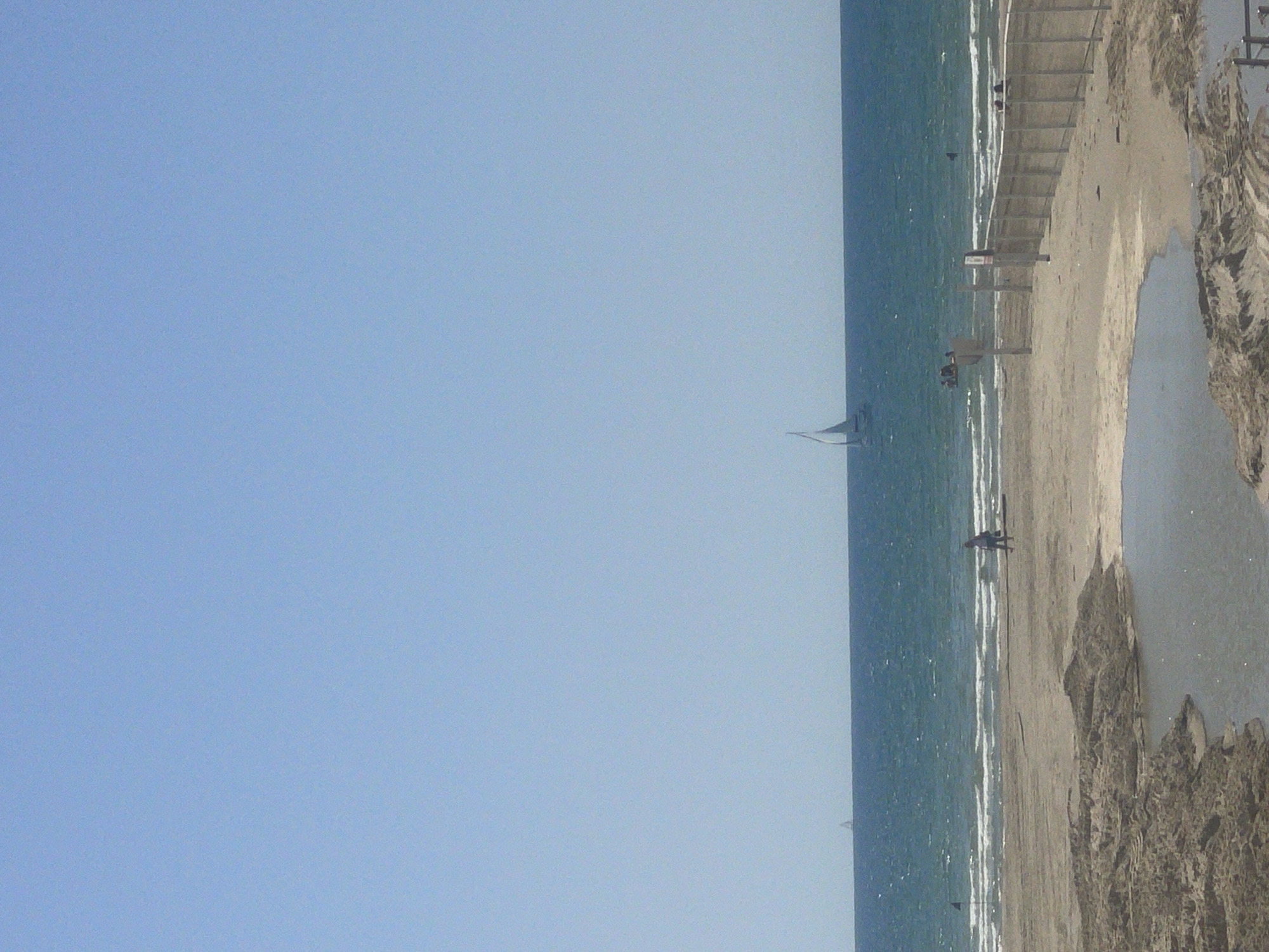 The image shows a beach scene in Herzliya, Israel. The ocean stretches horizontally, with a single sailboat visible in the distance. The beach is populated with people, some walking along the shore, while others are near a wooden structure that might be a lifeguard stand. A pool of water or large puddle is on the sand, creating a reflective surface. The sky above is clear and light blue.