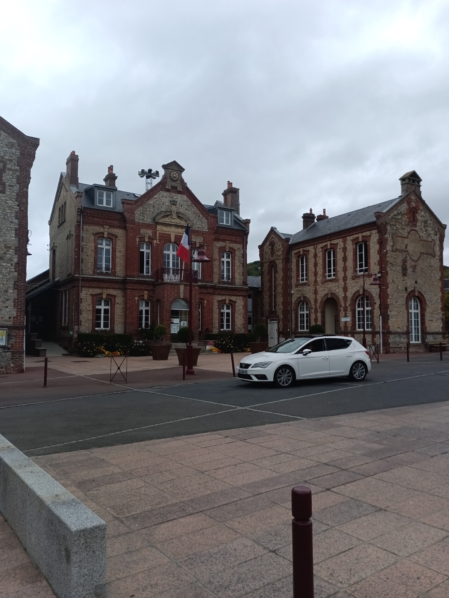 This is a medium shot of the Hôtel de Ville (Town Hall) in Houlgate, France, on an overcast day. A white hatchback car, possibly a Seat Leon, is parked in the foreground on the left side of the road, facing right. The car has a European license plate. Behind the car, the Hôtel de Ville is a two-story building constructed of red brick and stone, with a prominent French flag displayed on a flagpole to the left of the central entrance. The building has multiple windows with white frames, some arched. To the right of the Hôtel de Ville, another building of similar architectural style, also made of brick and stone, stands. The area in front of the buildings is paved with cobblestones and has some planters with yellow flowers. A lamppost is visible in front of the Hôtel de Ville. The sky is cloudy and grey.