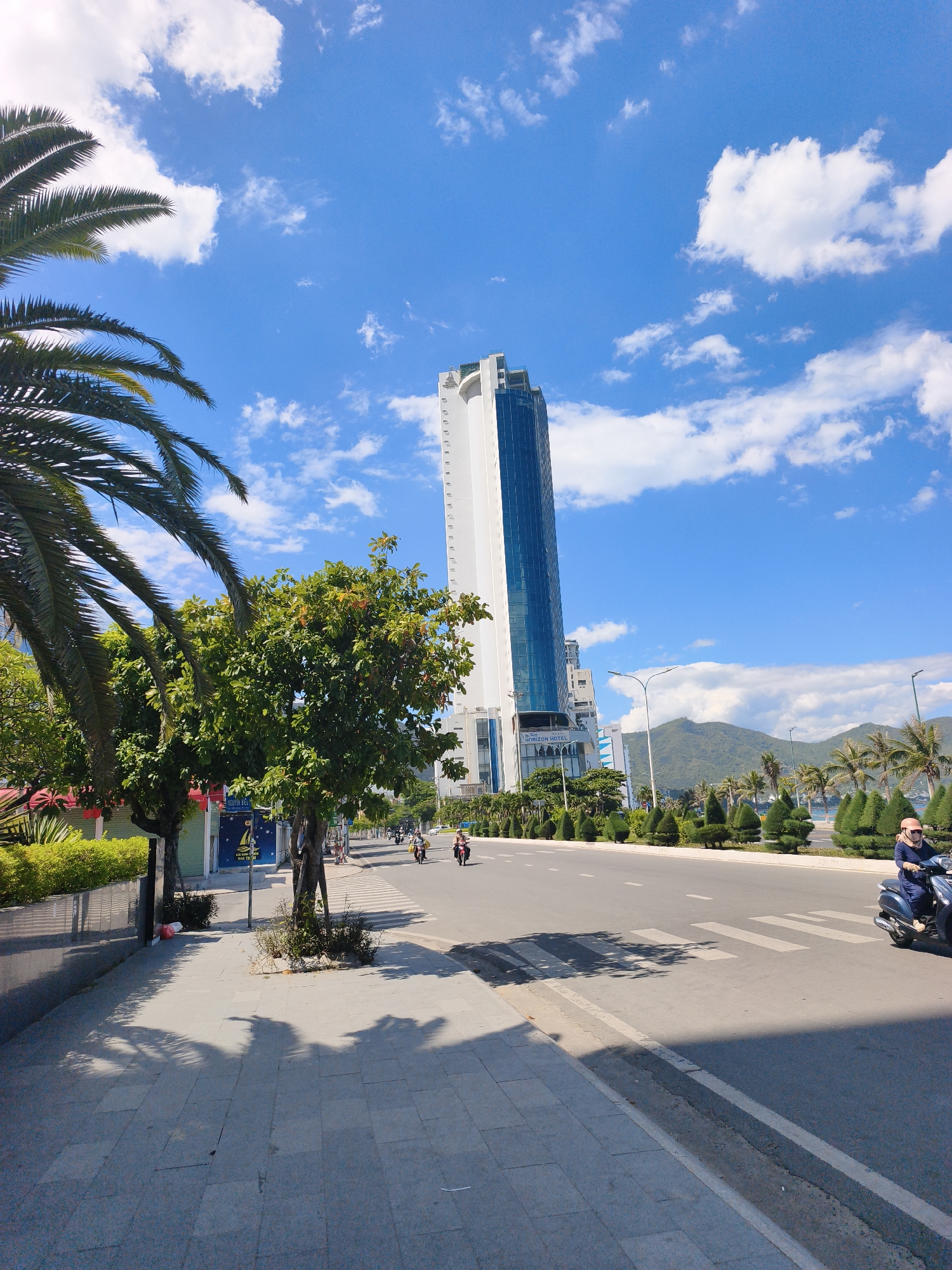 This is a street-level, wide-angle shot of a city scene in Nha Trang, Vietnam, on a bright, sunny day with blue skies and scattered white clouds. The perspective is from a sidewalk looking towards a multi-lane road with light traffic.
In the foreground, a paved sidewalk with gray square tiles is visible, casting distinct palm tree shadows from the left. A dark gray drainage channel runs along the edge of the sidewalk.
The road features a white pedestrian crossing. On the road, two motorcycles are present. The motorcycle on the right is ridden by a person wearing a blue outfit and an orange helmet. Further down the road, a few other motorcycles are seen with riders.
Behind the road, there's a neatly landscaped area with small, conical evergreen trees and palm trees. Beyond this, a tall, modern white skyscraper with blue-tinted windows dominates the middle ground. The building has a distinctive geometric design at its top. To the left of the skyscraper, a leafy green tree stands. On the far left, a large palm frond extends into the frame from the top.
In the background, a mountain range is visible. The overall impression is a coastal city environment with a blend of natural elements and modern architecture.