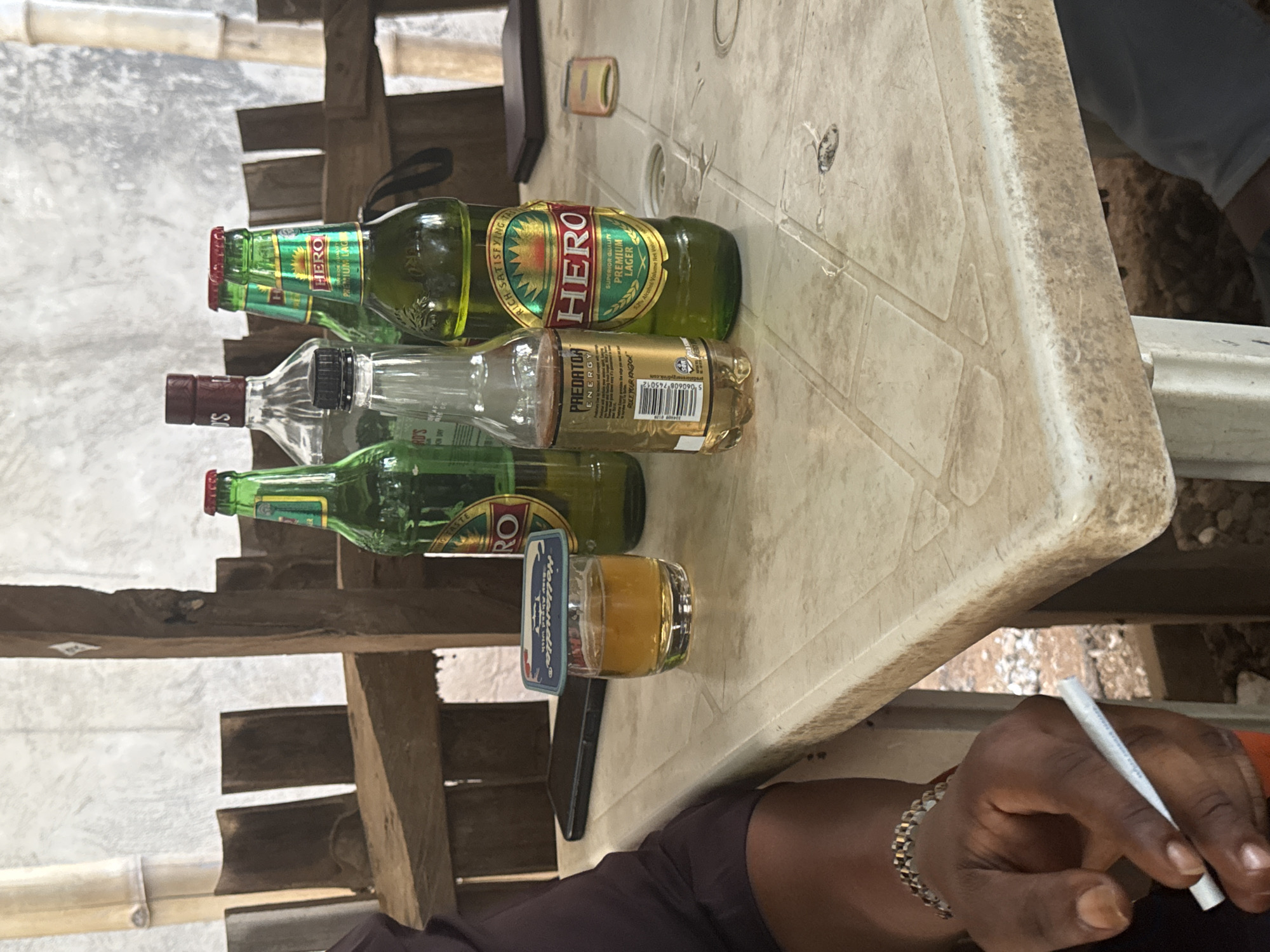The image captures a casual scene featuring multiple beverages on a worn, light-colored plastic table, likely in an outdoor or semi-outdoor setting in Kurunduma, Nigeria.
The main subjects are several bottles and a glass. Two green bottles of "HERO" beer are visible; one appears mostly full, while the other is partially consumed. The labels clearly display "HERO", "SUPERIOR QUALITY", "PREMIUM LAGER", and "RICH SATISFYING TASTE". Next to these is a clear, empty glass bottle. A golden-colored bottle of "PREDATOR ENERGY" drink, with the slogan "RULE YOUR KINGDOM", is also present. A small glass containing an amber liquid, likely an alcoholic beverage, rests on a coaster.
In the foreground, a dark-skinned hand, adorned with a silver-colored chain bracelet, is holding a lit cigarette. A dark object, possibly a mobile phone or a lighter, is visible near the glass. The table itself appears well-used and slightly dirty. The background consists of various wooden planks and bamboo structures, suggesting a simple, rustic environment. The overall scene depicts a moment of relaxation or a social gathering involving drinking and smoking. No specific time of day or weather conditions can be determined from the image.