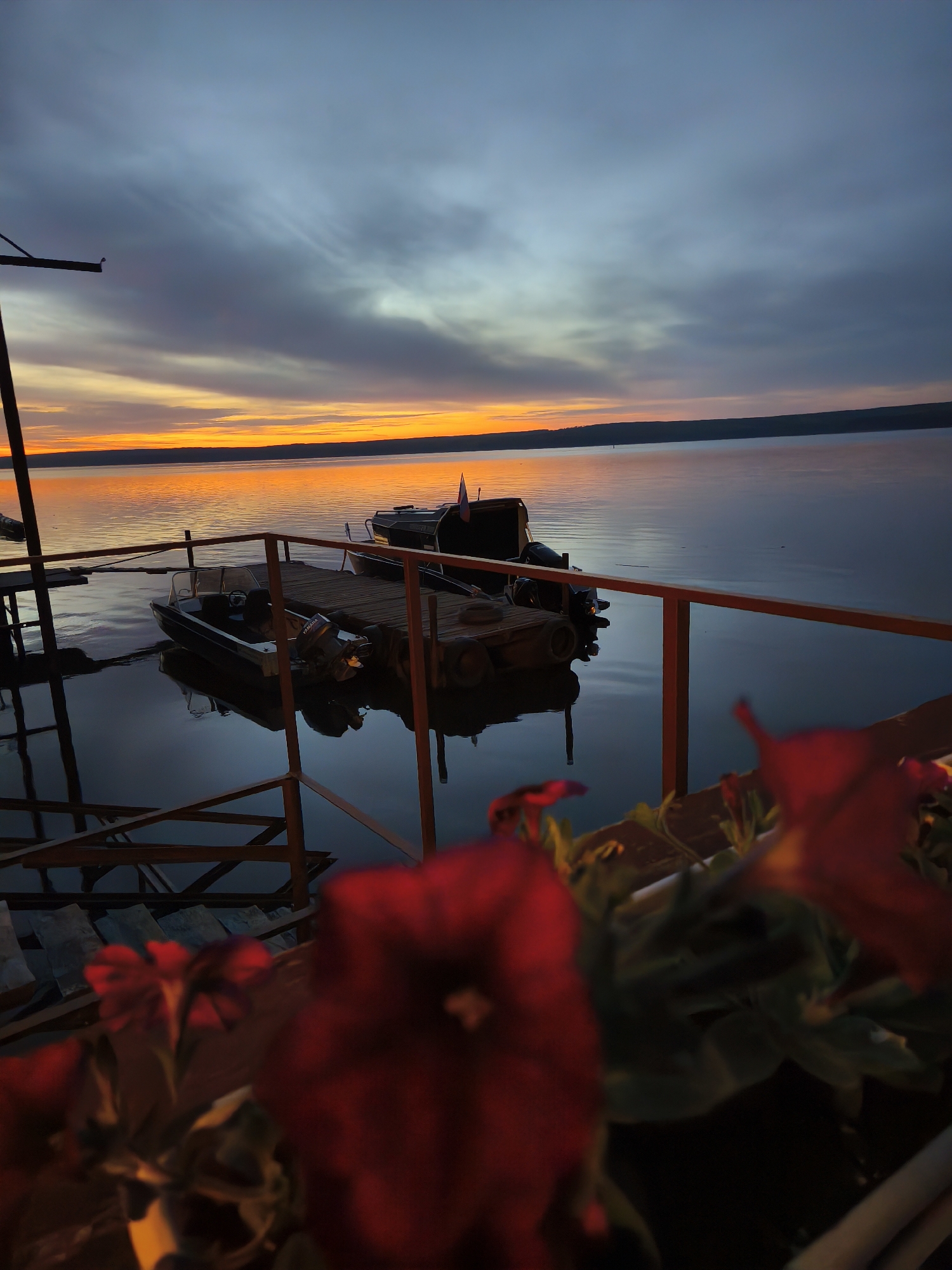 A scenic shot taken at dusk in Chaikovsky, Russia, portrays a tranquil waterfront scene. Two boats are moored at a wooden dock, with one displaying a small Russian flag. The sky displays a vibrant orange sunset, gradually transitioning into a darker, clouded blue. The calm water reflects the sky's colors and the outlines of the boats. In the foreground, out-of-focus red flowers frame the view, adding a touch of color contrast. A part of the dock's railing and support structures are visible. The distant shoreline is dimly lit, suggesting a hilly or forested landscape.