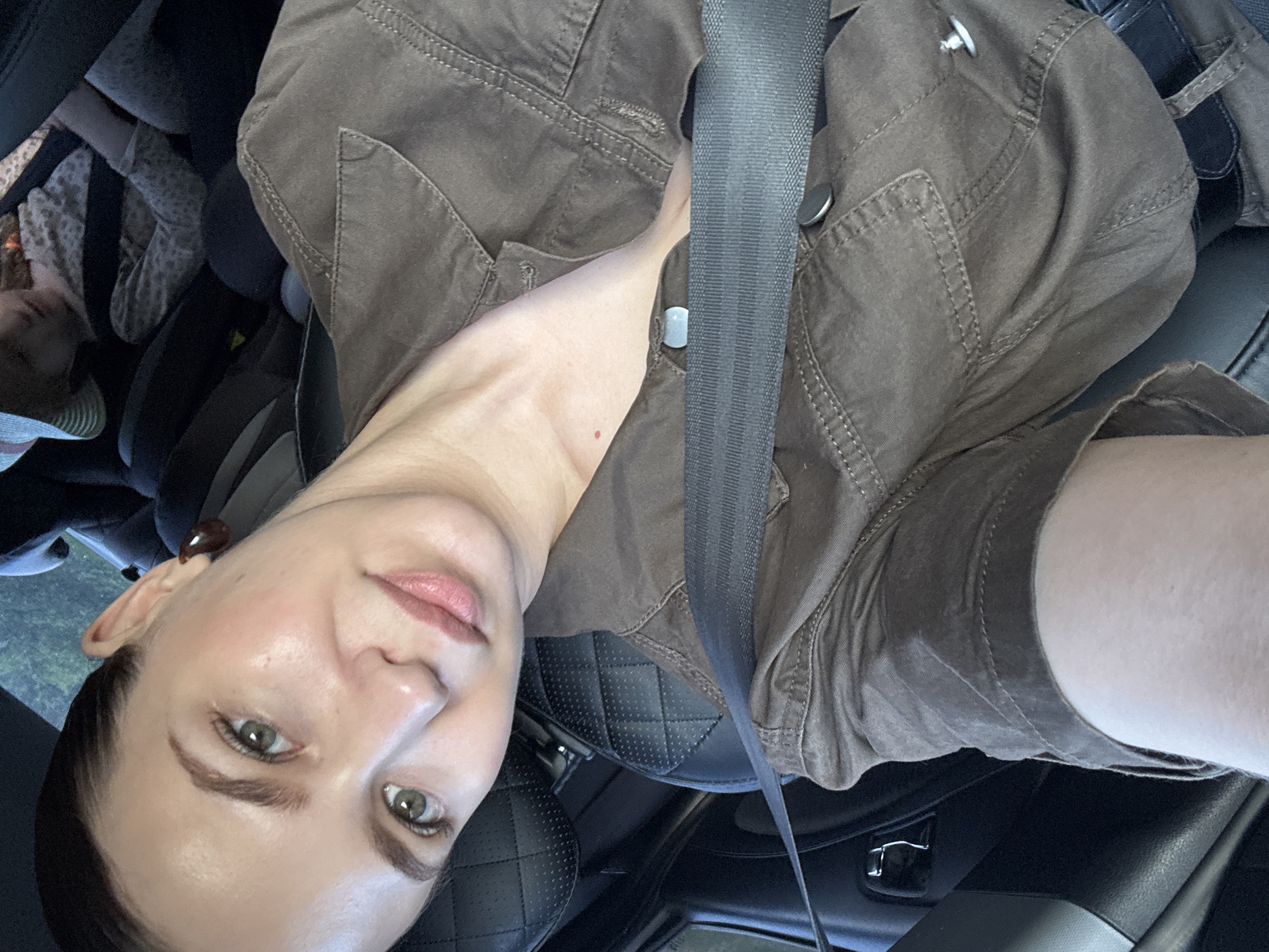 An adult woman is shown in a close-up selfie taken from a low angle inside a vehicle. She is wearing a brown button-up shirt and has her seatbelt fastened across her chest. Her head is tilted, and she is looking towards the camera. Her left eye is in focus, and she appears to have light makeup on. Her hair is pulled back. In the background, a child is visible in a car seat. The interior of the car includes black leather seats and a gray seatbelt. The lighting suggests it is daytime. The scene is not identifiable as being located in the city of Chaikovsky, Russia.