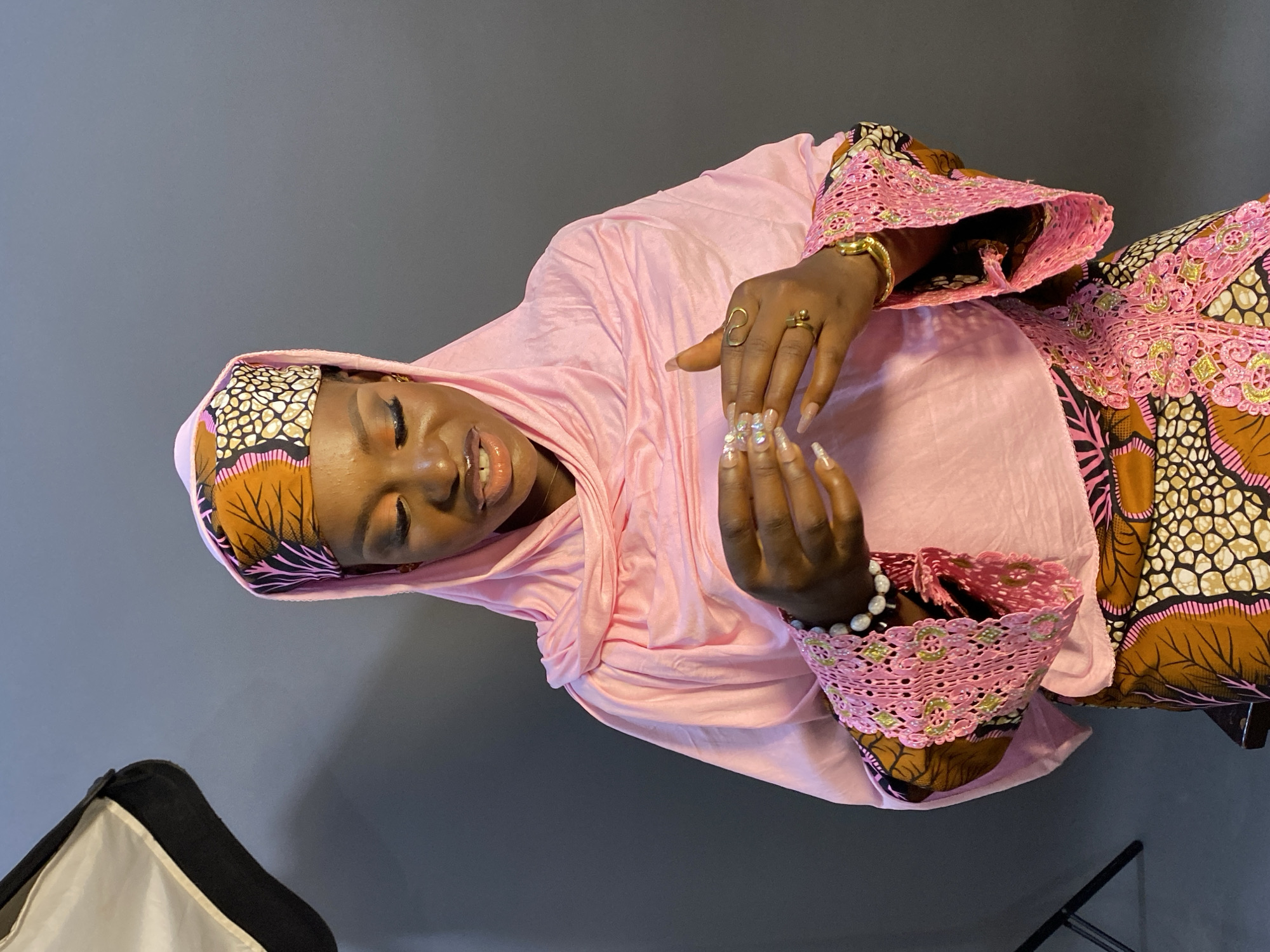 The image features a young Black woman posing for a photograph against a plain, dark grey backdrop, indicating an indoor studio setting. The photograph is rotated 90 degrees counter-clockwise.
The woman wears a soft pink hijab or headscarf that drapes gracefully around her head and shoulders. A patterned band in shades of brown, black, white, and pink is visible underneath the hijab, framing her face. She is dressed in a loose-fitting, long-sleeved garment, likely a dress or top, which is also pink. Beneath this, an elaborate patterned fabric, mirroring the colors of her headscarf band, is visible, particularly on her sleeves, which are further embellished with intricate pink lace and subtle gold-colored accents.
Her eyes are closed, and she has a gentle, serene smile, conveying a sense of peace and contentment. Her hands are held together in front of her, palms slightly open, in a delicate pose. Her long, artificial nails are clear or light-colored and adorned with shimmering decorations or gems. She wears gold-toned rings on her index and middle fingers and a white pearl-like bracelet on her right wrist.
In the bottom left corner, part of a white softbox, a piece of photography lighting equipment, is visible. A black stand is also partially seen in the bottom right. The overall impression is a graceful and culturally rich portrait from Jos, Nigeria. No visible text is present in the image.