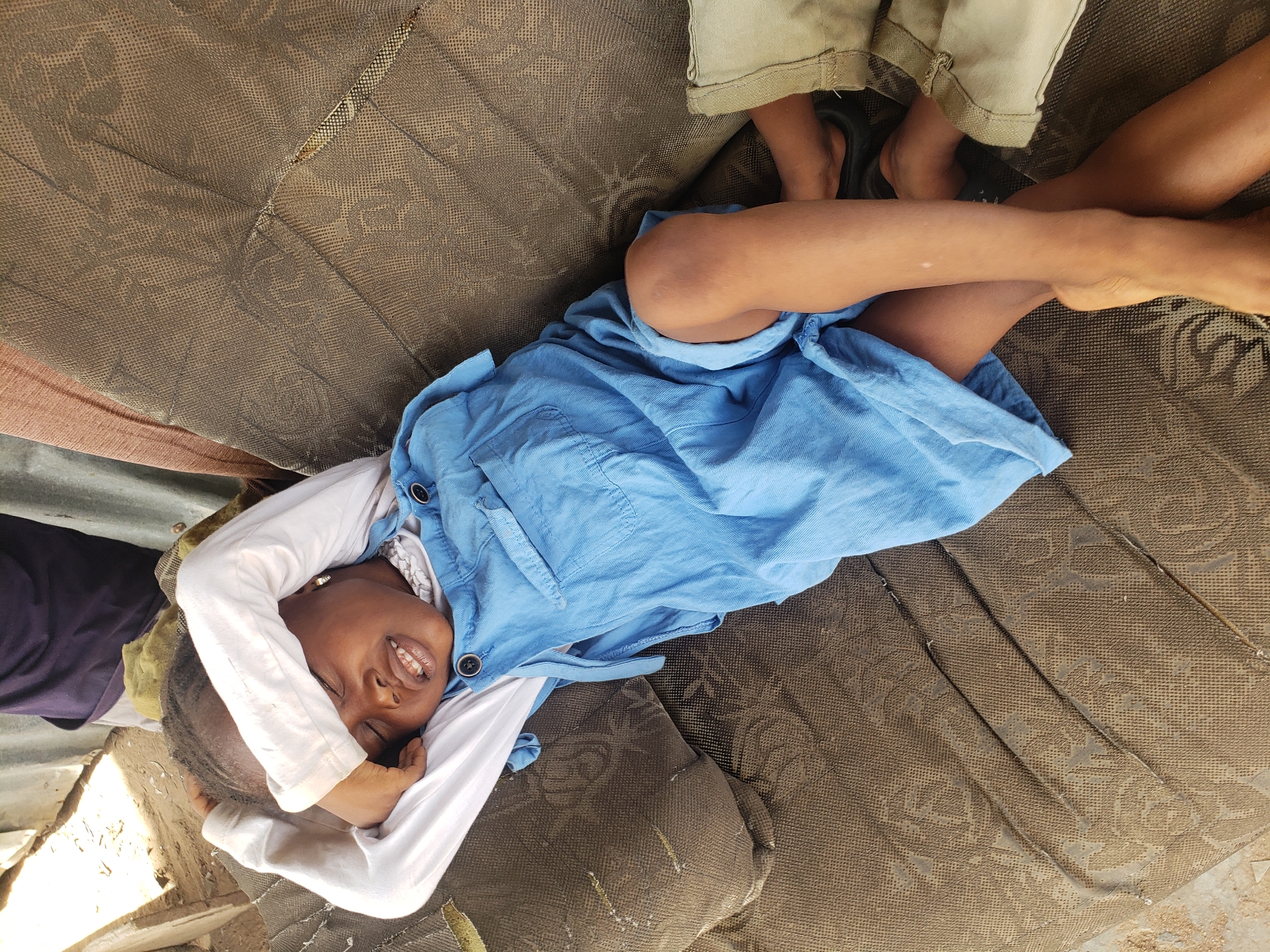 A young child, likely a girl, is prominently featured in this image from Adankolo, Nigeria, lying on their back and smiling brightly at the camera. The child wears a light blue sleeveless dress over a white long-sleeved shirt, with hands casually placed behind their head or covering part of their face. Their eyes are mostly closed in a joyful, squinting smile, revealing their teeth.
The child is resting on a worn, brown patterned mattress or padded surface, which shows signs of age and use. In the upper right corner, the lower legs and feet of another person are visible, clad in light-colored shorts or trousers and dark slippers, suggesting companionship or close proximity. To the far left, a dark purple fabric and glimpses of what appears to be corrugated metal and uneven ground are visible, indicating a humble, possibly outdoor or very simple indoor setting.
The bright, clear lighting suggests it is daytime, likely sunny, contributing to the child's squinting smile. The overall emotion conveyed by the child is one of happiness and contentment. There is no visible text in the image.