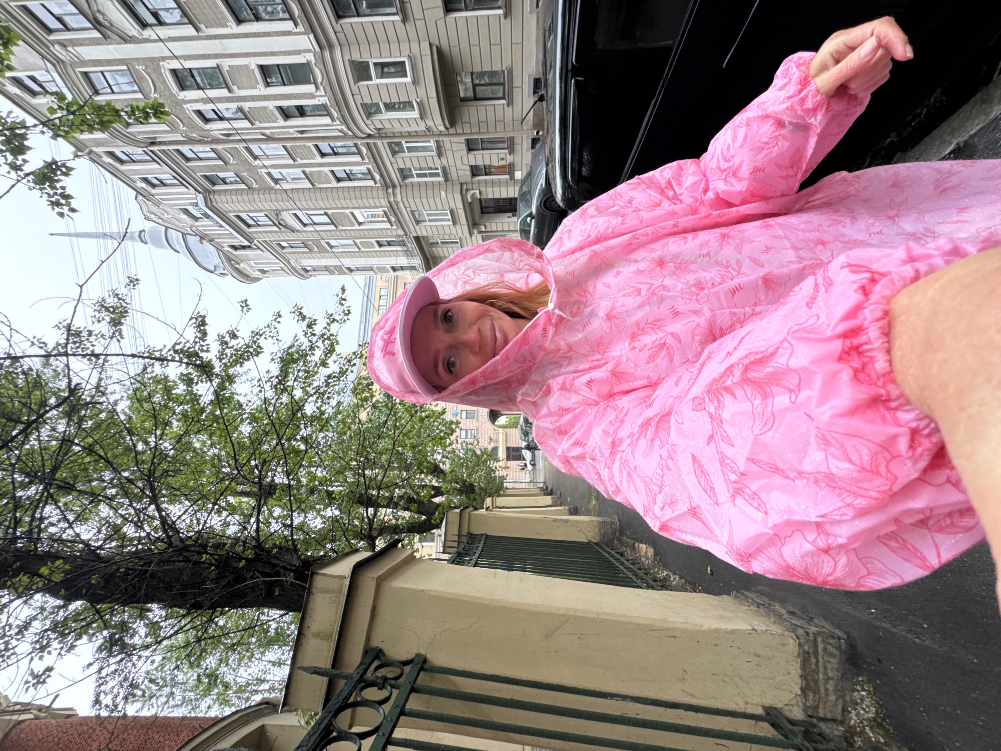 A woman wearing a pink rain poncho and matching hat stands on a sidewalk in Saint Petersburg, Russia. The perspective is rotated, placing the building on the left. The building is multi-storied with light-colored walls and many windows. The sidewalk is adjacent to a fence made of a tan-colored wall and a dark green iron grate. Trees line the street. In the background, a tall, thin spire is visible, characteristic of Saint Petersburg's architecture. A black car is parked on the right side of the frame. The scene appears to be taken on a rainy day.