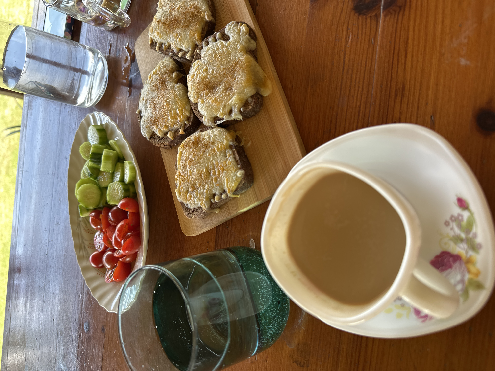 A table scene depicts a meal set up. On the table, there are slices of bread topped with melted cheese arranged on a small wooden cutting board. A small dish contains sliced cucumbers and halved tomatoes. A teacup filled with a light brown liquid sits on a floral-patterned saucer. Two glasses, one empty and one appearing to contain water, are also present. The table appears to be made of wood. The scene is illuminated by natural light, possibly from a nearby window. There is no indication of Lehchevo, Bulgaria, in the image.