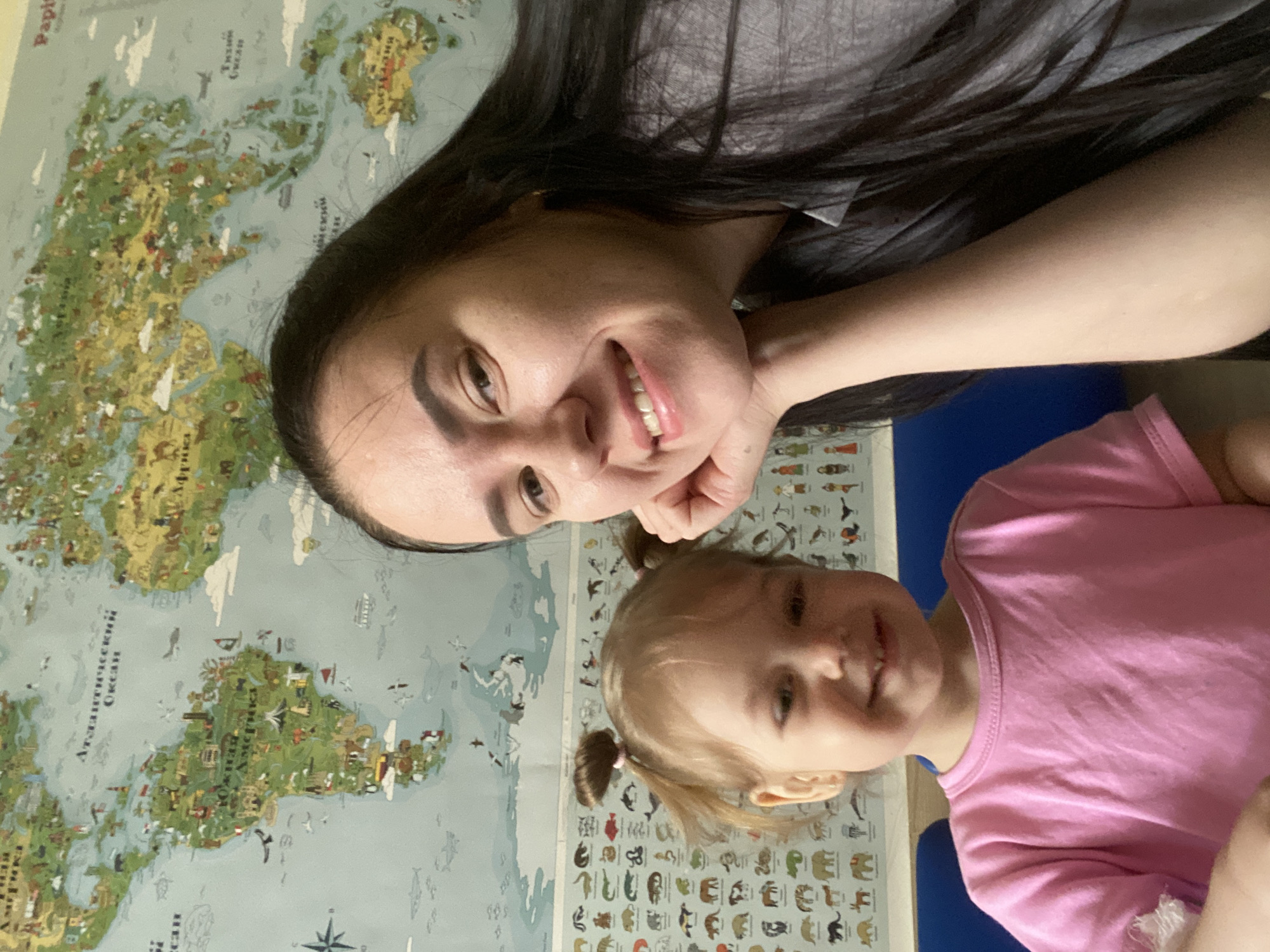 A woman and a young child are visible in the image. The woman has long, dark hair and is smiling. The child, wearing a pink shirt, has fair hair styled in pigtails. They appear to be posing for a photo in front of a wall with a map of the world and a section with pictures of animals. The location of this image within the city of Perm, Russia, cannot be determined from the provided visual information.