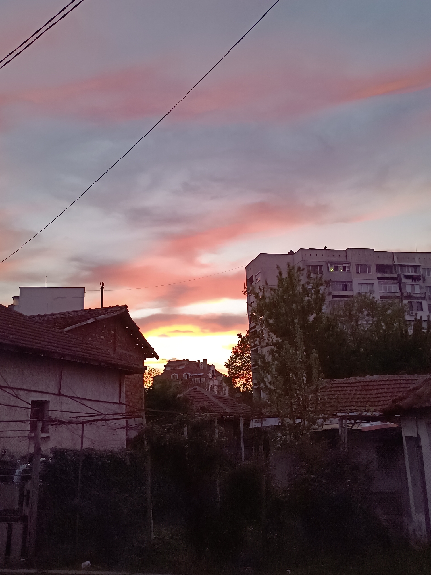 The image displays an urban or semi-urban landscape during a vibrant sunset. The sky is the dominant feature, filled with clouds painted in brilliant shades of pink, orange, and yellow near the horizon, gradually fading into purple and grey higher up. Two dark power lines diagonally cross the upper portion of the sky.
In the foreground, to the left, an older house with a reddish-brown tiled roof and light-colored walls is visible, accompanied by a white, flat-roofed structure behind it. A slender chimney-like feature extends from the roofline. To the right, more tiled roofs and structures are partially obscured by dense vegetation and fencing.
In the mid-ground, a large, multi-story apartment block with a grey facade and numerous windows stands out against the colorful sky. Trees and bushes, appearing as dark silhouettes, are scattered throughout the scene, especially in front of and around the buildings. Another building with distinctive rounded windows and a dark roof is faintly visible in the distance, nestled among the trees.
The scene is captured at sunset, indicated by the intense, low-angle light and the dramatic cloud colors. No specific activities are depicted. The blend of older, tiled-roof houses and the more modern apartment block suggests a residential area, possibly in an Eastern European context. There is no visible text in the image. The overall impression is one of natural beauty contrasting with an everyday urban setting.