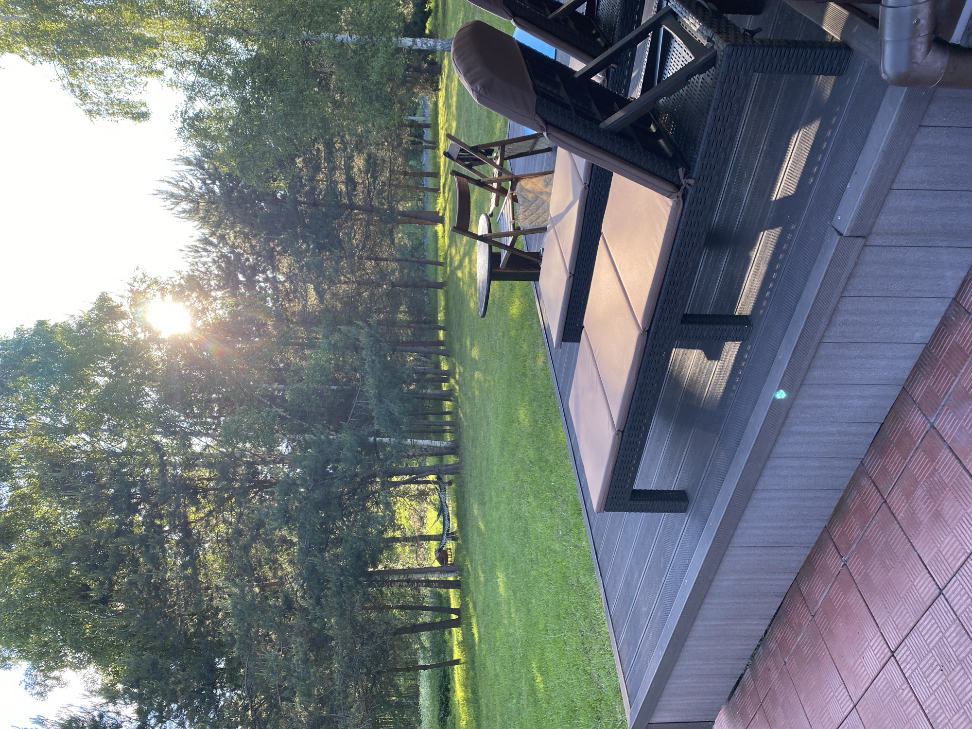 This image, captured in Копнино, Russia, presents an outdoor scene featuring a deck, lawn, and surrounding forest under bright, low-angle sunlight. In the foreground, a dark gray composite deck occupies the lower half of the frame. Its side is clad with vertical gray panels, and a small green light is visible on the right side panel. A portion of a red-brown tiled surface with a textured pattern is present in the bottom-left corner, adjacent to the deck.
On the deck, two dark synthetic wicker sun loungers with light brown cushions are positioned towards the right, oriented away from the viewer. To their left, a dark round table is accompanied by two folded wooden chairs. A light-colored, possibly quilted, bag hangs on the back of the left wooden chair.
Beyond the deck, a manicured green grass lawn stretches into the midground. The lawn is bordered by a dense line of trees, including numerous tall, slender coniferous trees with dark green needles and lighter-barked deciduous trees with lighter green foliage. A green and black striped hammock is suspended between two trees in the mid-left section of this treeline.
The background is filled with the upper canopy of the forest. The sun is visible in the upper-center-left, emitting a strong lens flare and creating bright overexposed areas in the visible sky, which appears light blue or white. The lighting casts long, distinct shadows across the deck and lawn. No individuals are present in the scene.