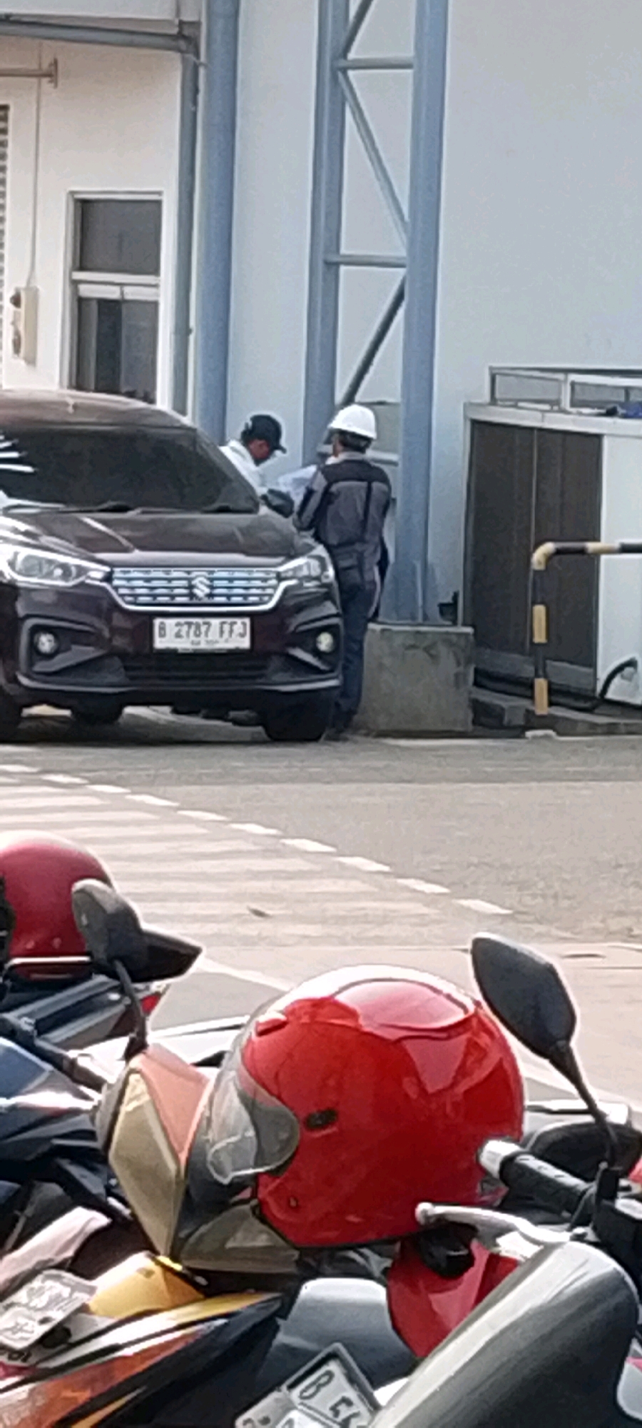 In the city of Depok, Indonesia, at the entrance of a facility, two individuals are seen interacting near a dark maroon Suzuki Ertiga with license plate B 2787 FFJ. The person on the left, wearing a dark baseball cap and a light-colored shirt, appears to be looking at or discussing something with the person on the right. The individual on the right is wearing a grey work uniform and a white hard hat, holding what looks like a document or blueprint. In the foreground, several red and yellow motorcycle helmets are visible, along with parts of motorcycles. The background shows a portion of a building with white walls and large industrial air conditioning units.