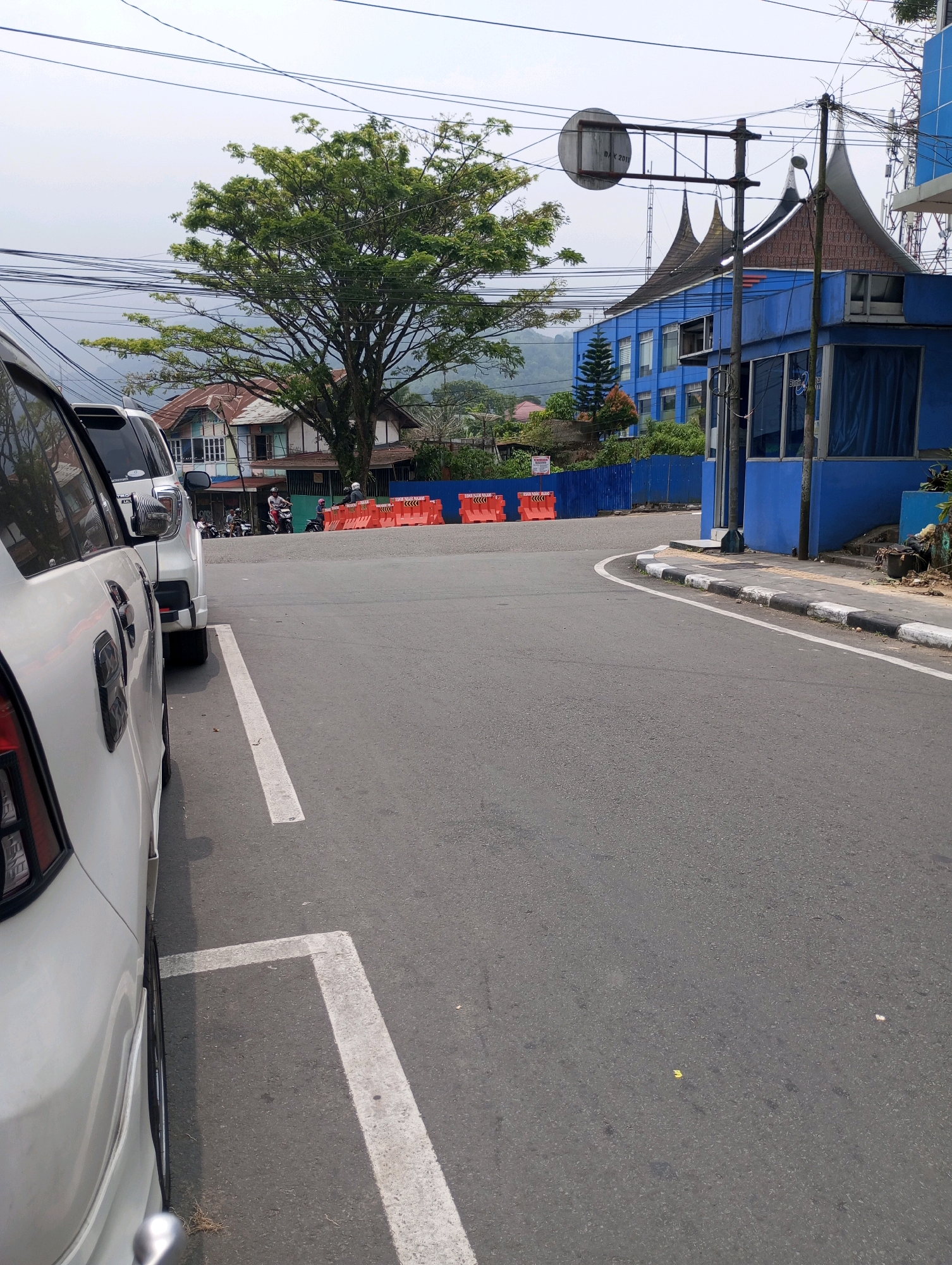 In Padang Panjang, Indonesia, a street scene is captured on an overcast day. In the foreground, the side of a white car is visible, parked on the left side of the frame, partially obscuring a white parking line. The car's door handle and a portion of its rear light are discernible. The asphalt road curves towards the right, marked by a white line. A sidewalk with black and white painted curbs runs along the right side of the road.
Further down the road, several bright orange traffic barriers are placed across what appears to be a construction or road closure zone. Beyond the barriers, a traditional Indonesian house with a distinctive multi-tiered roof and white-paneled walls stands under a large tree with lush green foliage.
To the right of the road, a modern blue building is prominent, featuring glass windows and a blue fence. A street light and electrical poles with numerous wires crisscross the sky. In the background, more buildings and the hilly landscape typical of the Padang Panjang region are visible under a hazy sky. A few individuals on motorcycles are seen in the middle ground near the house. The overall impression is a moderately busy urban environment with a blend of traditional and modern architecture, amidst a backdrop of natural hills.