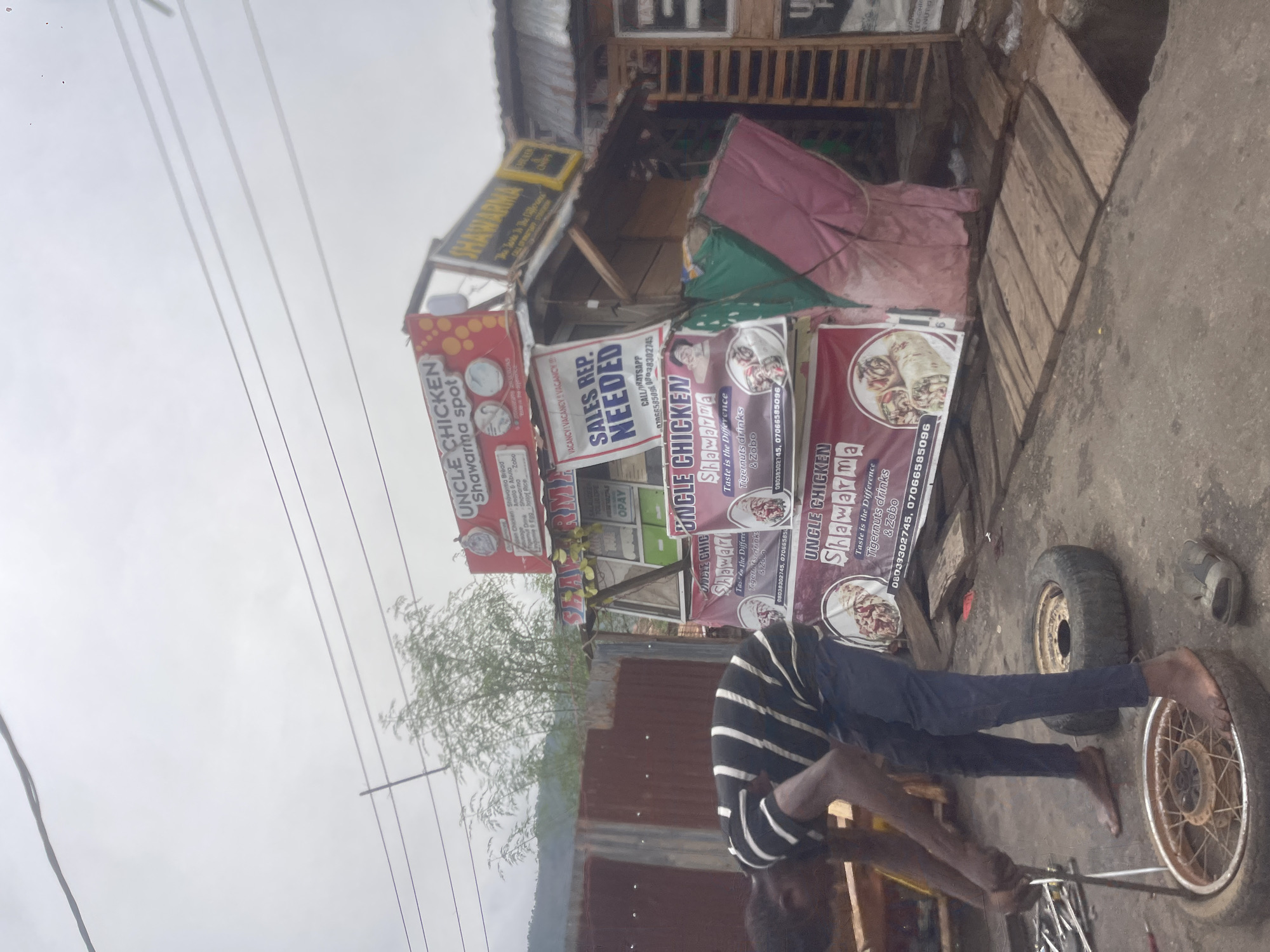 The image captures an outdoor scene in Lokoja, Nigeria. In the foreground, a man in a black and white striped shirt and dark pants is seen working on a bicycle tire. He is barefoot and appears to be using tools to repair the tire. Several tires are visible around him, indicating a possible roadside repair activity. In the background, there is a food stall or a small eatery named "Uncle Chicken Shawarma Spot." The stall has multiple signs advertising shawarma, other food items, and drinks. A sign also indicates a vacancy for sales representatives. The surroundings seem urban, with a mix of temporary structures and perhaps residential buildings in the distance. The sky is overcast, suggesting a cloudy day.