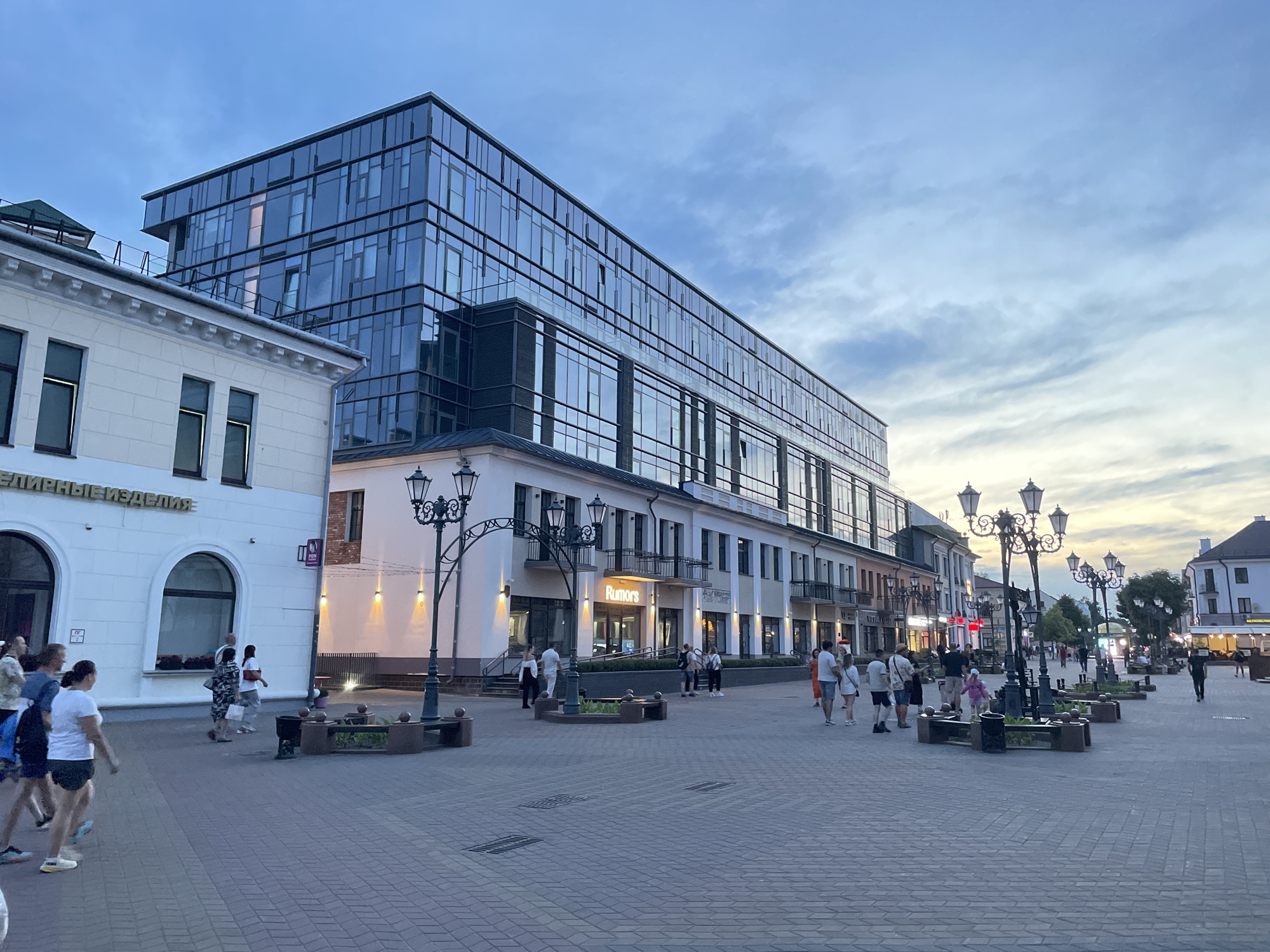 This is a wide-angle, eye-level outdoor photograph of a pedestrian street in Brest, Belarus, during twilight. The scene depicts a city street lined with buildings of varying architectural styles. On the left, a white, two-story building with arched windows and signage reading "единые изделия" (meaning "unified products" in Russian) is visible. Adjacent to it is a modern, multi-story glass-fronted building reflecting the evening sky.
The street is paved with red brick, and several ornate, black lampposts with multiple lights are positioned along the walkway. Benches with planters are situated between the lampposts. People are walking along the street, some singly, some in small groups. One building has a sign that reads "Rumors." The sky above is a mix of blue and grey clouds, with a hint of yellow and orange from the setting sun near the horizon on the right side of the frame. The overall atmosphere is calm and leisurely, typical of an early evening in a European city.