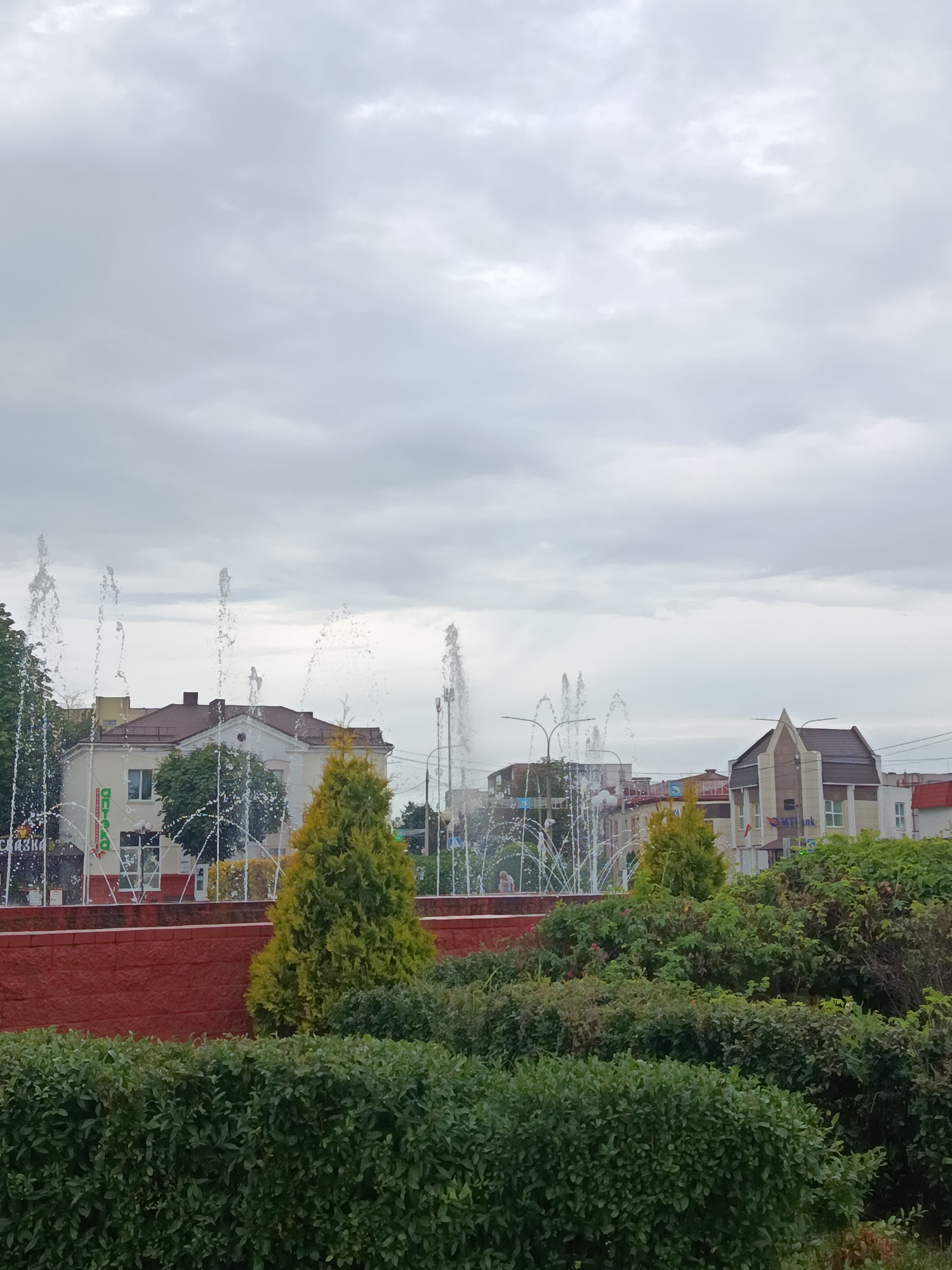 The image shows a cloudy sky over a city scene in Zhlobin, Belarus. In the foreground, there is a well-maintained hedge of green bushes. Behind the hedge, a red brick wall is visible, with a conical green tree planted on top. Further back, a series of fountains spray water upwards, creating a mist. Beyond the fountains, several buildings are visible, including a light-colored building with a brown roof and a statue on top, and another building with a darker, angular roofline featuring the logo of "MTBank". The general environment appears to be an urban park or public square, with a mix of greenery and architectural structures.