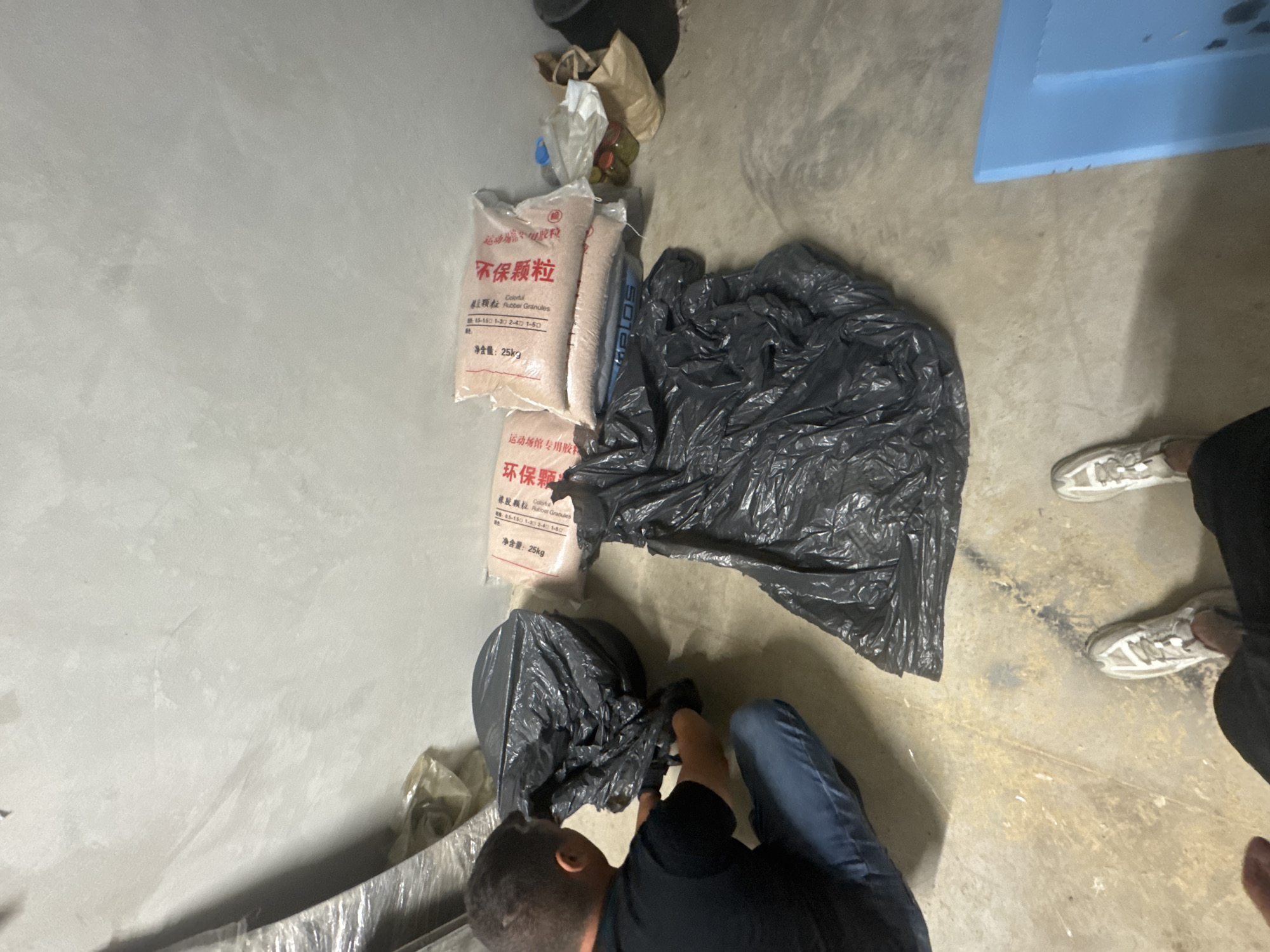 The image shows a person kneeling on a concrete floor next to several bags and a large crumpled black plastic bag. Two bags are labeled with Chinese characters and the words "Colorful Rubber Granules," indicating they contain rubber granules, likely for sports fields. The label specifies "25kg" as the net weight. Other items include additional bags and what appears to be packaging material. The person is wearing blue jeans, a black shirt, and white sneakers. Part of a blue container is visible in the upper right corner. The environment appears to be a storage or work area. The location of Sofia, Bulgaria is not discernible from the image.