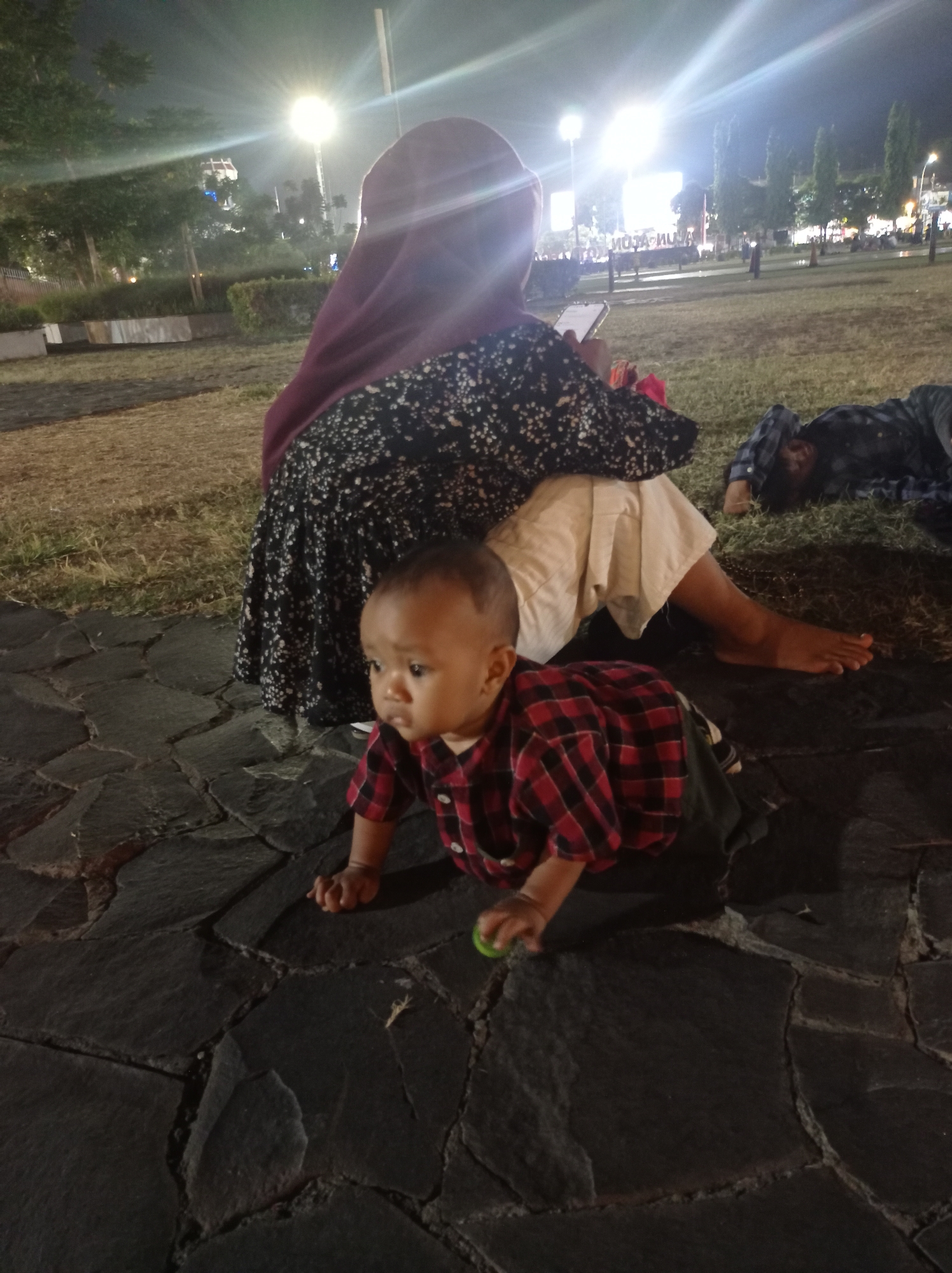 The image captures an outdoor scene at night, likely in Tegal, Indonesia, given the context. In the foreground, a baby is crawling on a paved area made of dark, irregularly shaped stones. The baby is wearing a red and black plaid shirt and appears to be looking towards the left of the frame. Behind the baby, a person wearing a dark hijab and a patterned dress is seated on the ground. Their legs are visible, with one foot bare. The person is holding a mobile phone in their hand. To the right and further back, another person is lying down on the grass, wearing a dark, checkered shirt. The background shows a park-like setting with trees, grass, and lighting fixtures that illuminate the area. In the distance, there are more trees and structures, and a large, illuminated banner or screen is visible. The overall impression is of a relaxed evening gathering in a public outdoor space.