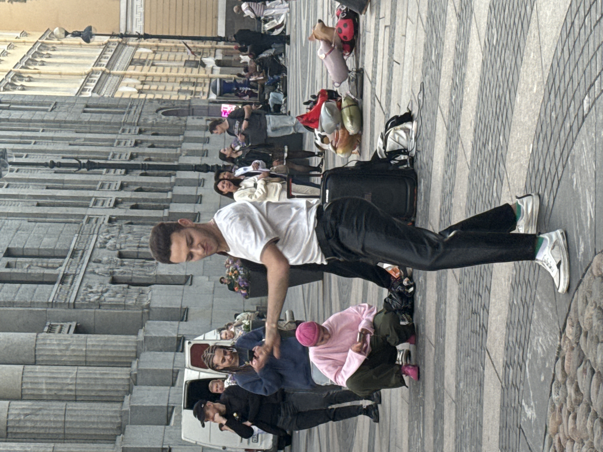 The image depicts a street scene in Saint Petersburg, Russia, likely on Nevsky Prospekt, a prominent avenue known for its grand architecture and bustling atmosphere. The architecture visible suggests a classical style with tall, fluted columns and ornate detailing on the building facades.
In the foreground, a young man with short, dark, curly hair is captured mid-dance. He is wearing a white t-shirt and black, loose-fitting pants, with white sneakers and green socks. His body is turned to the side, with his left arm extended, as if reaching out or in motion. He appears to be the central focus of the scene, perhaps performing for onlookers.
Behind him, a person wearing a pink beanie and a light pink oversized sweatshirt is crouched down, looking at a mobile phone. They are wearing dark green pants and bright pink sneakers. To the left, several other individuals are visible. One person is wearing a black baseball cap and dark clothing, seemingly observing the dancer. Further back, a woman with braided hair is also looking at her phone.
Scattered on the pavement are several inflatable objects, including what appears to be a ladybug, a pig, and possibly a cow-patterned balloon, along with some bags and a dark suitcase. These items suggest a public space where people might be gathering or perhaps a street vendor's setup. The ground is paved with large, rectangular grey stones interspersed with lighter grey grout lines, forming a patterned walkway. In the background, more people are milling about, some seated and others standing, contributing to the sense of a lively urban environment. A white van is parked on the left side of the image. The overall lighting suggests an overcast day, with diffused light casting soft shadows.