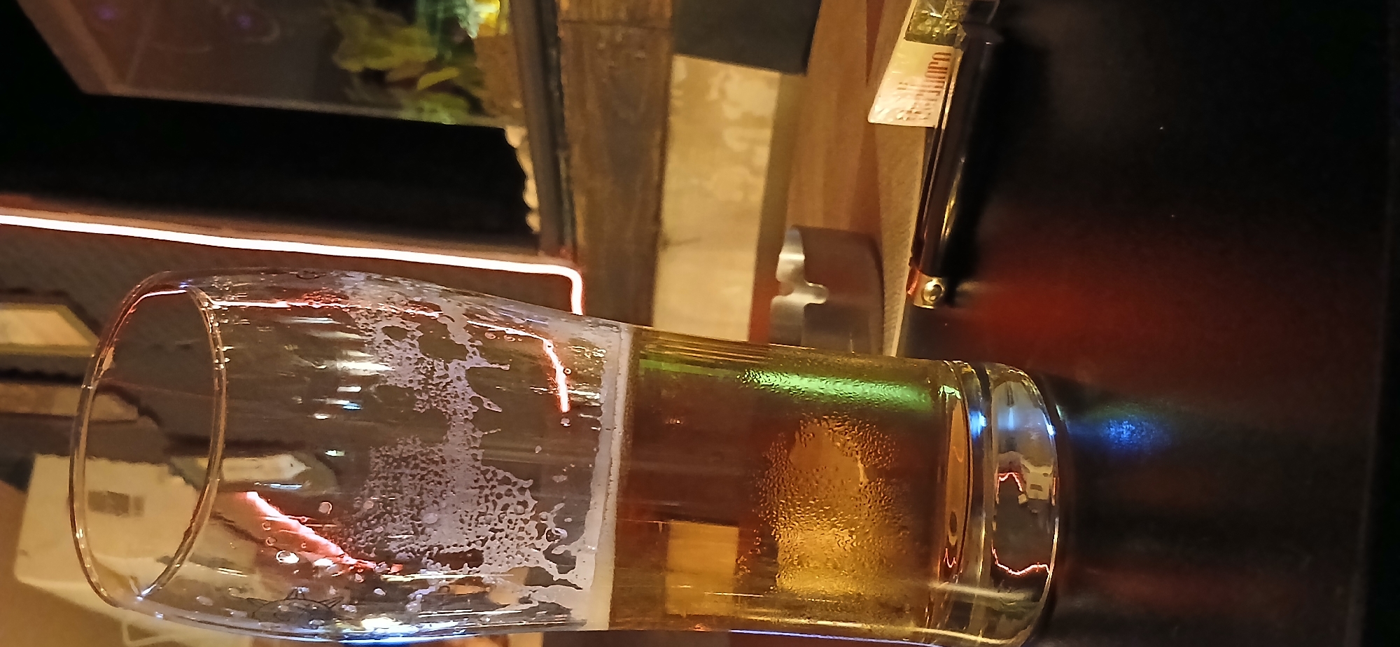 A close-up, low-angle shot captures a partially full pint glass of beer, condensation beading on its surface, resting on a dark, reflective tabletop. The beer is golden in color with a frothy head near the rim. To the right of the glass, a silver ashtray and a cigarette pack with the "Marlboro" logo are visible. A dark-colored lighter with a metallic tip lies next to the cigarette pack. In the background, out of focus, is a shelving unit with various items, including what appears to be a small aquarium with green plants and pebbles. The scene is dimly lit, with warm-toned lights casting reflections on the glass and tabletop, and a neon light strip running vertically in the background. The overall setting suggests an indoor environment, possibly a bar or pub. There are no people visible in the image. The specific location in Yerevan, Armenia, cannot be determined from the visual information provided.