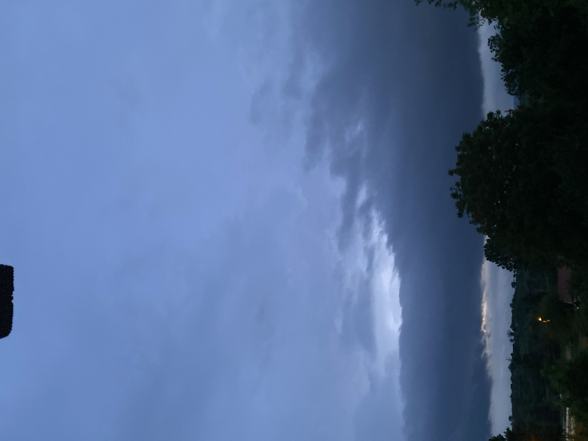 The image shows a sky dominated by dark, ominous clouds. The lower right corner contains trees and a hill, indicating an outdoor scene. The trees are dark green, and the hill appears to have some buildings, one with a light. The sky is mostly dark gray, suggesting stormy weather. The location is likely in a rural or suburban area, potentially near Konstantinovo, Bulgaria, given the user's prompt. No people are visible. The overall environment appears to be on the verge of a heavy downpour.