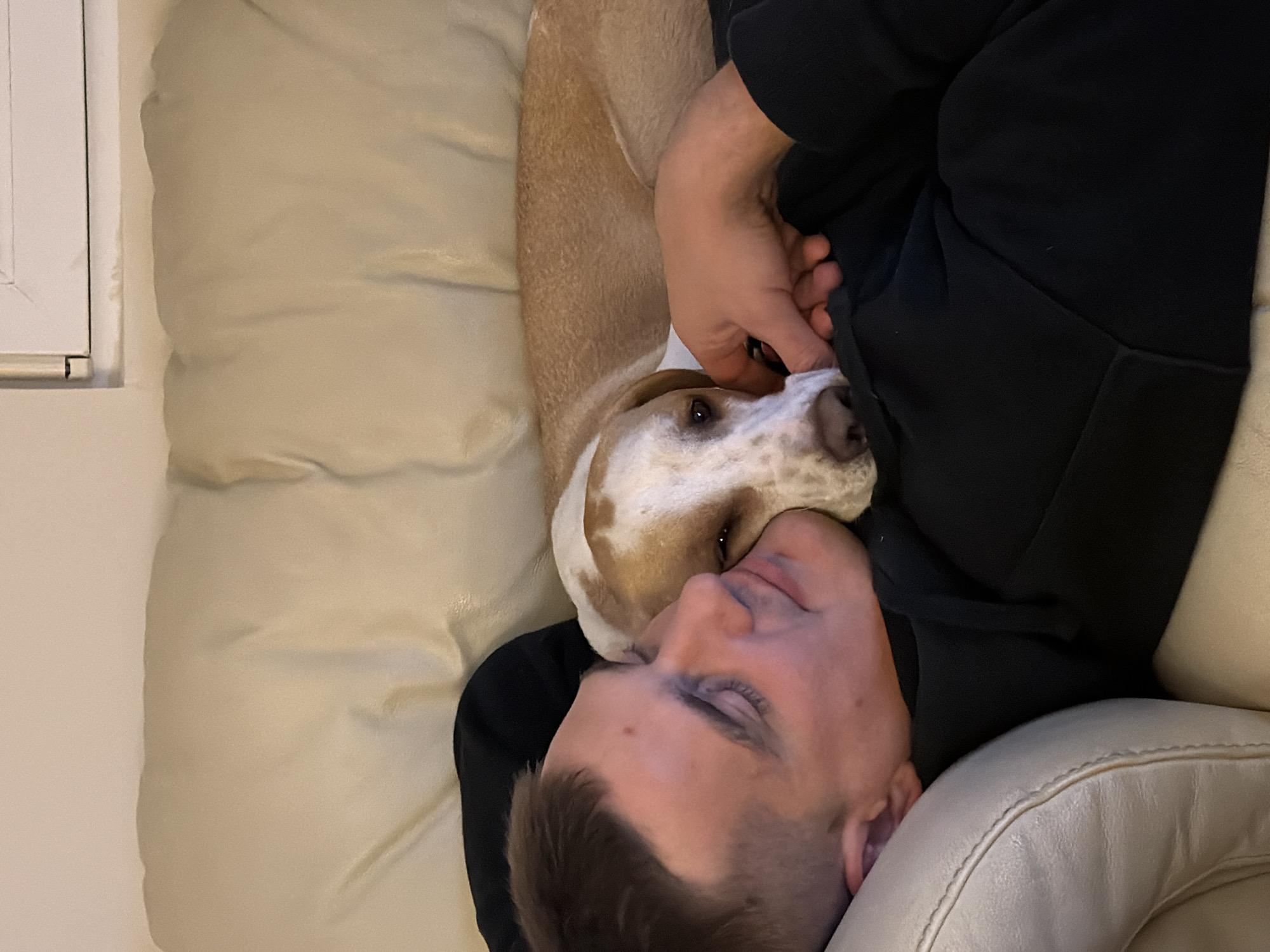 A man is lying on a white leather couch with a tan and white dog resting its head on his chest. The man is wearing a black hoodie. A window or door frame is visible in the upper left corner of the image. The location is not identifiable as being in Budva, Montenegro.
