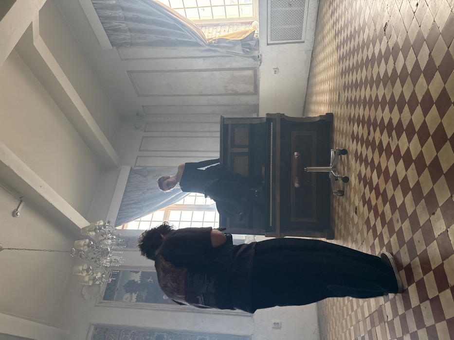 The image depicts an indoor scene, likely a photoshoot, within a large, high-ceilinged room in Moscow, Russia. In the foreground, a person with their back to the camera, wearing a dark t-shirt and dark trousers, is operating a camera or recording device. Centrally, a dark wooden upright piano serves as a prop. A woman, dressed in a black off-shoulder top and a long dark skirt, is seated atop the piano, posing with her head tilted slightly downwards. A silver-legged office-style swivel stool with a brown padded seat is positioned directly in front of the piano.
The room features a red and white checkered tile floor. The walls exhibit decorative moldings, with faded blue-toned murals or tapestries integrated into panels on the left side. A multi-branched crystal chandelier hangs from the ceiling in the upper-left quadrant. Multiple tall, multi-pane windows with light blue drapes line the background walls, admitting bright natural light that illuminates a visible atmospheric haze within the room. Radiator grates are present beneath the windows on the right. The environment appears aged, with noticeable wear on the walls and floor.