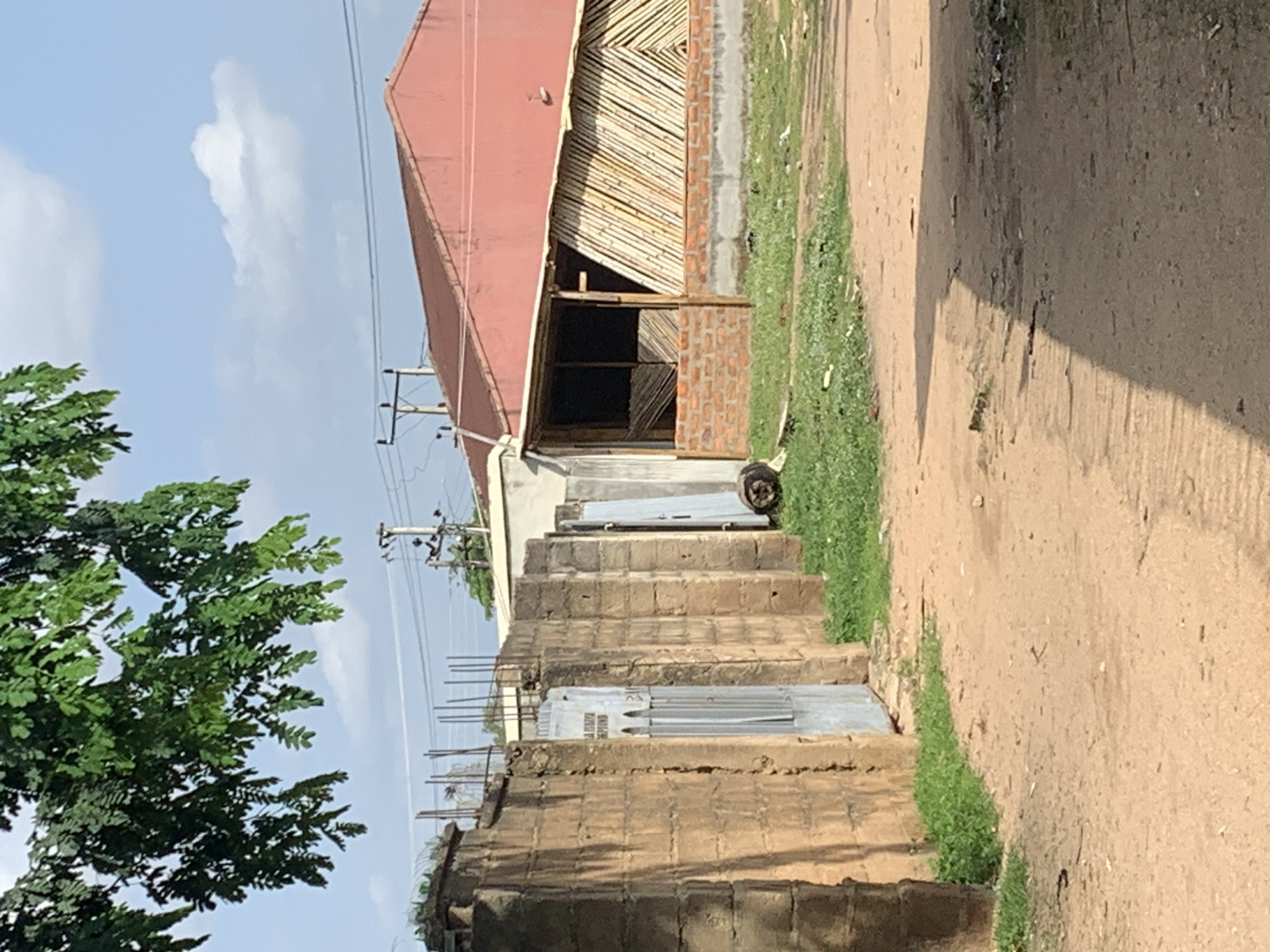 In an outdoor, daylight scene, a dirt road passes by a wall made of tan cinder blocks, with a rusty metal gate set into it. Beyond the wall is a building with a red roof and a structure made of bamboo poles. Green grass grows around the base of the building and along the wall. In the background, a tree with green leaves hangs over the scene, and telephone poles with wires are visible against a blue sky with scattered clouds. The overall impression is one of an undeveloped or rural area.