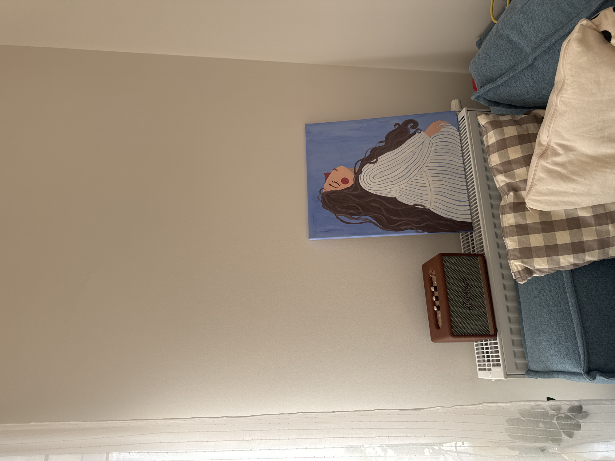A painting of a woman laying down with long brown hair on a blue background is hanging on a beige wall. Below the painting is a brown Marshall speaker mounted on a white radiator. A bed with blue and patterned pillows is situated next to the radiator. A white curtain is visible at the bottom of the image. There is no evidence to indicate this scene is in Люк, Russia.