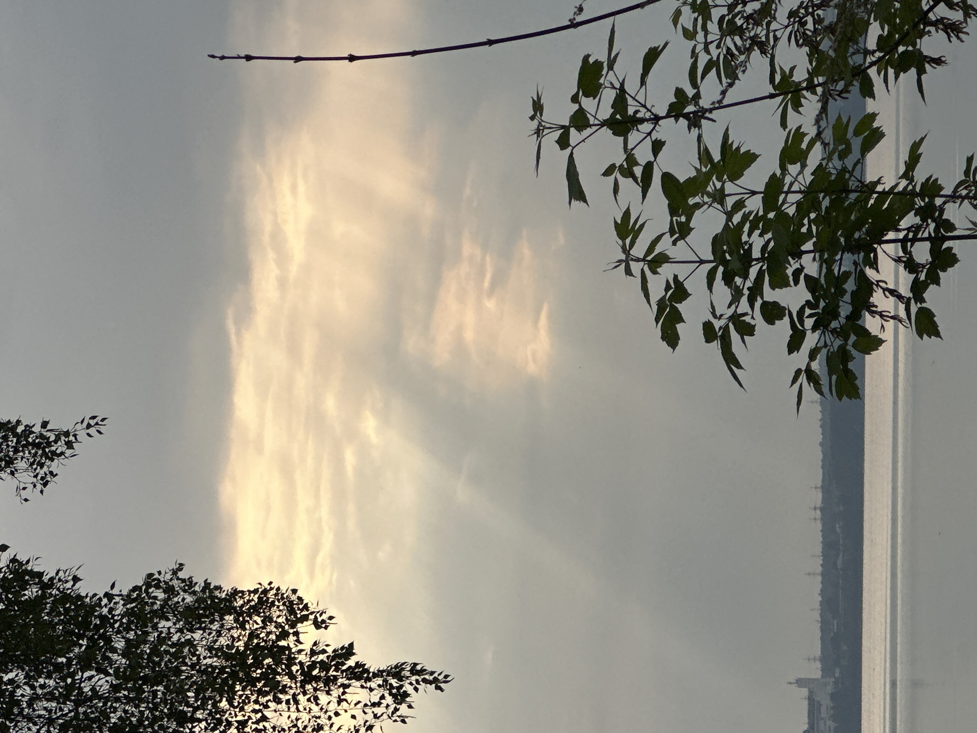 The image shows a body of water, likely a lake or river, with trees silhouetted in the foreground. The sky is overcast with visible light rays breaking through the clouds. A city skyline is barely visible on the opposite shore of the water. The location appears to be Исеть, Russia.