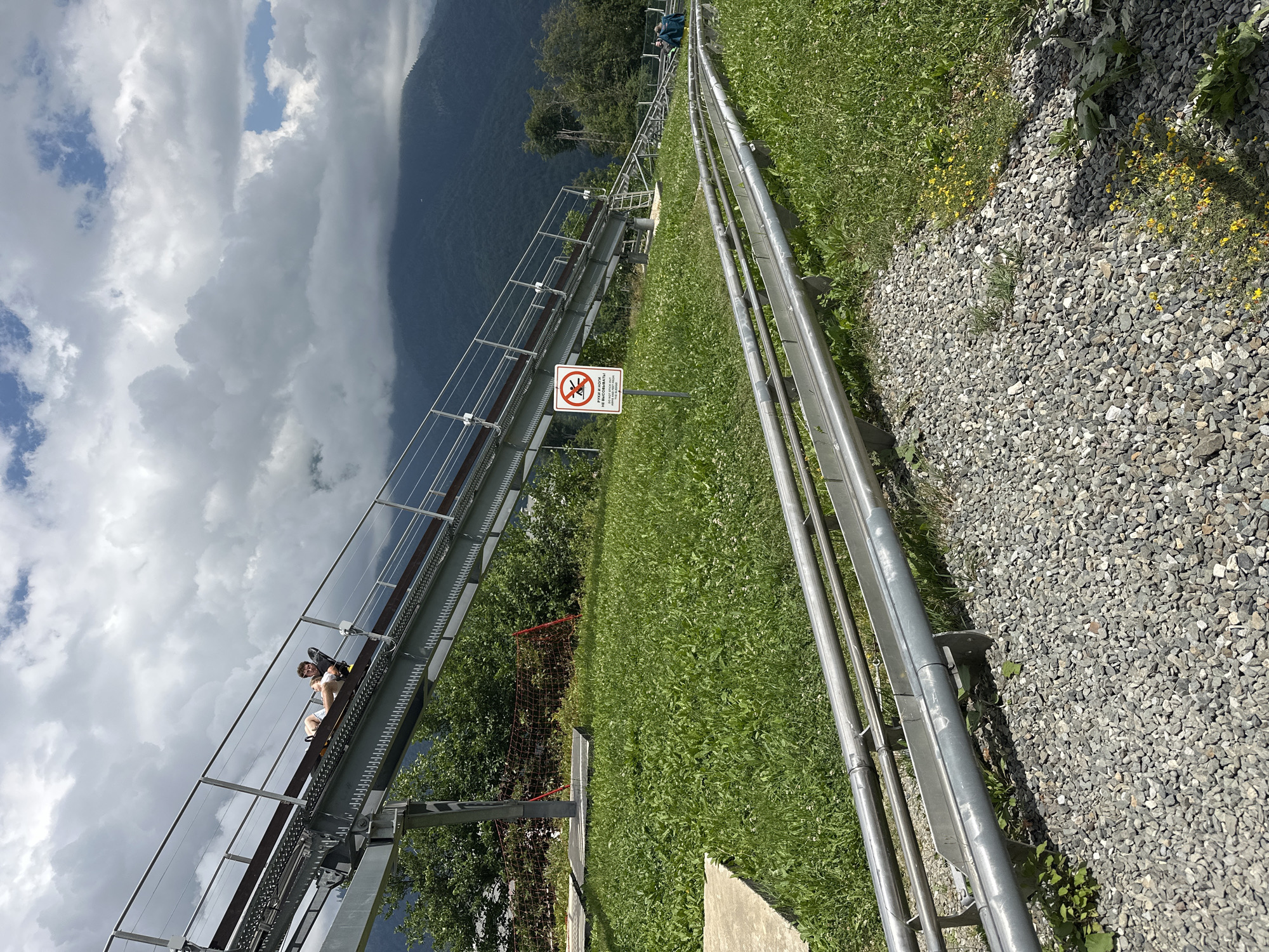 This is a full-shot, eye-level, outdoor photograph of a gravity roller coaster attraction in Estosadok, Russia. The attraction consists of a metal track elevated on a metal support structure, winding down a grassy slope. In the foreground, a section of the metal track is visible, laid on a bed of grey gravel. Further up the slope, the track ascends, with a person riding a cart on it. The person, a young man wearing a white t-shirt and grey shorts, is reclined in the cart, facing forward and looking to his left. He appears to be enjoying the ride. A warning sign with pictograms of a person with their arms and legs extended, and text in Russian, is positioned to the left of the track. The background features lush green vegetation, trees, and a misty mountain range under a cloudy sky. The overall impression is that of a recreational activity in a natural setting.