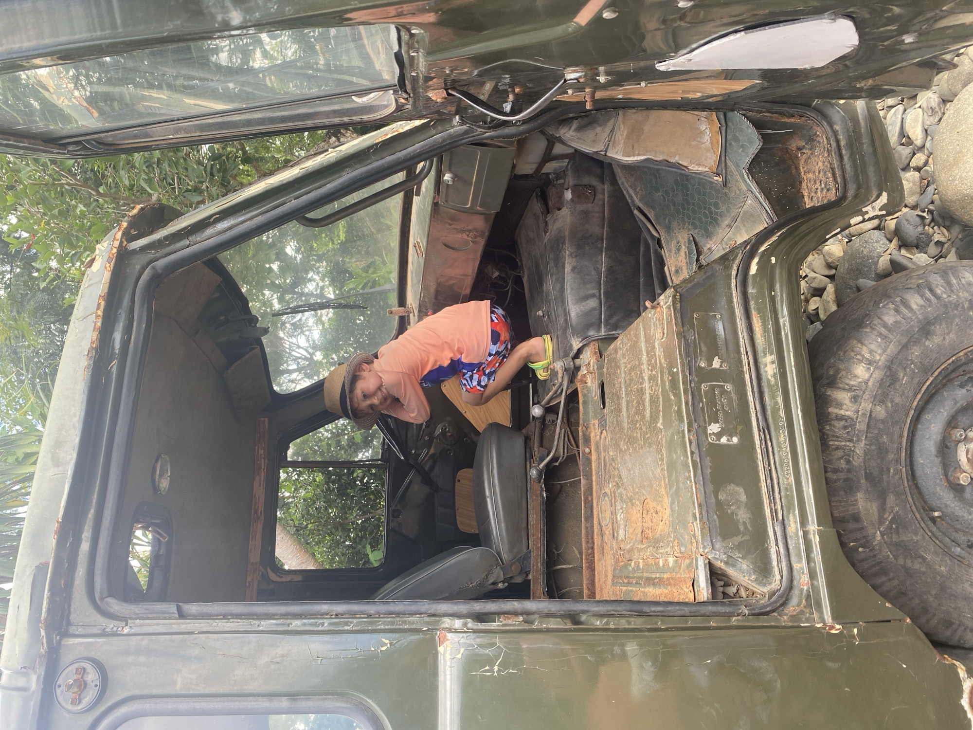 A young boy wearing a straw hat, an orange t-shirt, and patterned swim trunks squats inside the driver's side of a rusty, olive-green military truck. The truck appears to be abandoned and is parked on a rocky surface amidst lush green foliage. The boy is looking towards the camera with a smile, holding his hands near his face. The interior of the truck is worn and damaged, with exposed metal and peeling paint. The scene is set outdoors, with trees and vegetation visible in the background. The location is identified as Tien Thanh Ward, Vietnam.
