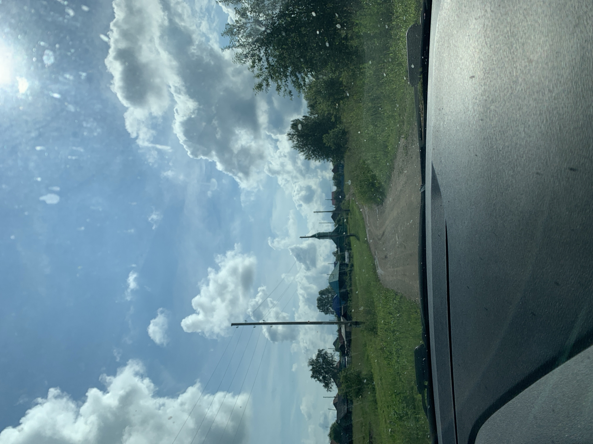 The image captures a view from inside a vehicle, looking out through a dusty windshield. A dirt road winds through a rural landscape with green grass and trees on either side. In the distance, a small village is visible with several houses and what appears to be a church with a steeple. Telephone poles and power lines run along the left side of the road. The sky is blue with large, fluffy white clouds. The overall impression is of a sunny day in a quiet, rural area, possibly in the city of Barsai, Russia, given the context.