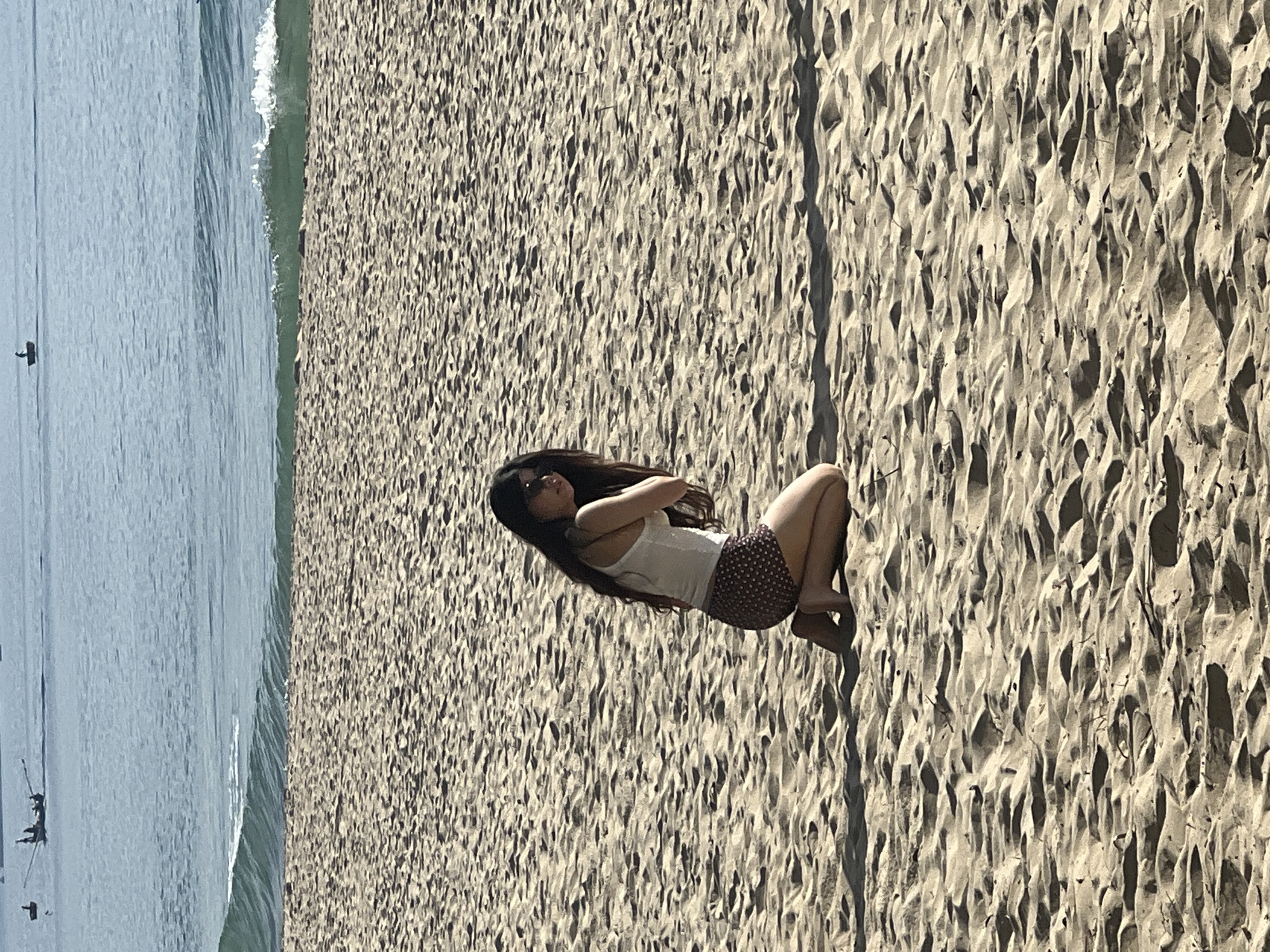 A young woman with long dark hair kneels on a sandy beach. She is wearing a white tank top and patterned shorts. Her body is turned away from the camera, and she appears to be looking down. The beach is covered in sand dunes, and the ocean is visible in the background. There are several small boats on the water. The scene appears to be shot during the daytime, with the sun casting shadows on the sand. This could be Mỹ Khê Beach in Đà Nẵng, Vietnam, known for its long stretches of sand and proximity to the sea.