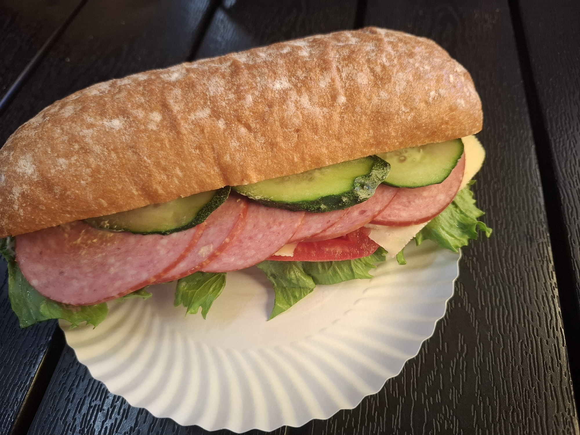 A close-up, high-angle shot shows a sandwich with salami, cucumber, lettuce, tomato, and cheese on a white paper plate. The sandwich is placed on a dark wooden table. The bread of the sandwich is a long, light brown roll with a dusting of flour. The salami slices are pink and arranged in layers. Sliced cucumbers are visible on top of the salami. Green lettuce leaves are peeking out from under the salami. A slice of red tomato is also visible. A slice of pale yellow cheese is under the salami and lettuce. The paper plate has a scalloped edge. The wooden table has a dark brown, textured surface. The scene is lit with artificial light, casting subtle shadows. There are no people or animals visible in the image. The location is Dubăsarii Vechi, Moldova.