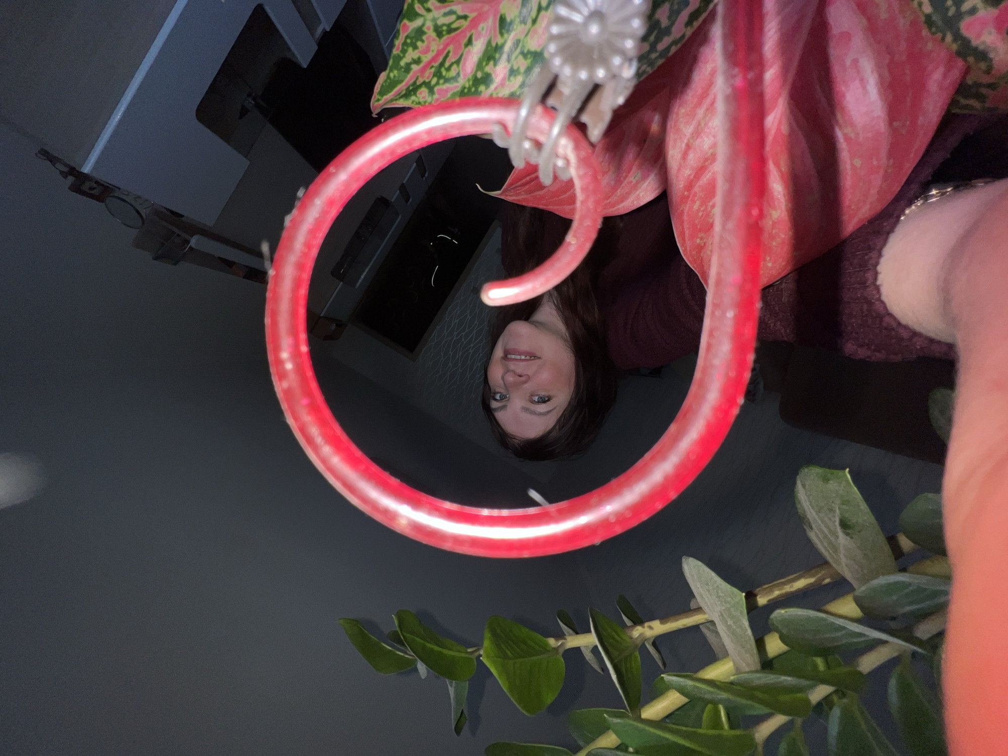A selfie taken indoors shows a woman's face centered within a red ring light. The woman has dark hair and light-colored eyes. Above and to the right of the ring light is a houseplant with pink and green leaves. Below the ring light is another houseplant with green leaves. Part of the woman's arm is visible in the upper right corner of the image. A cabinet and other blurred objects are visible in the background. There is no specific information in the image that identifies the location as Perm, Russia.