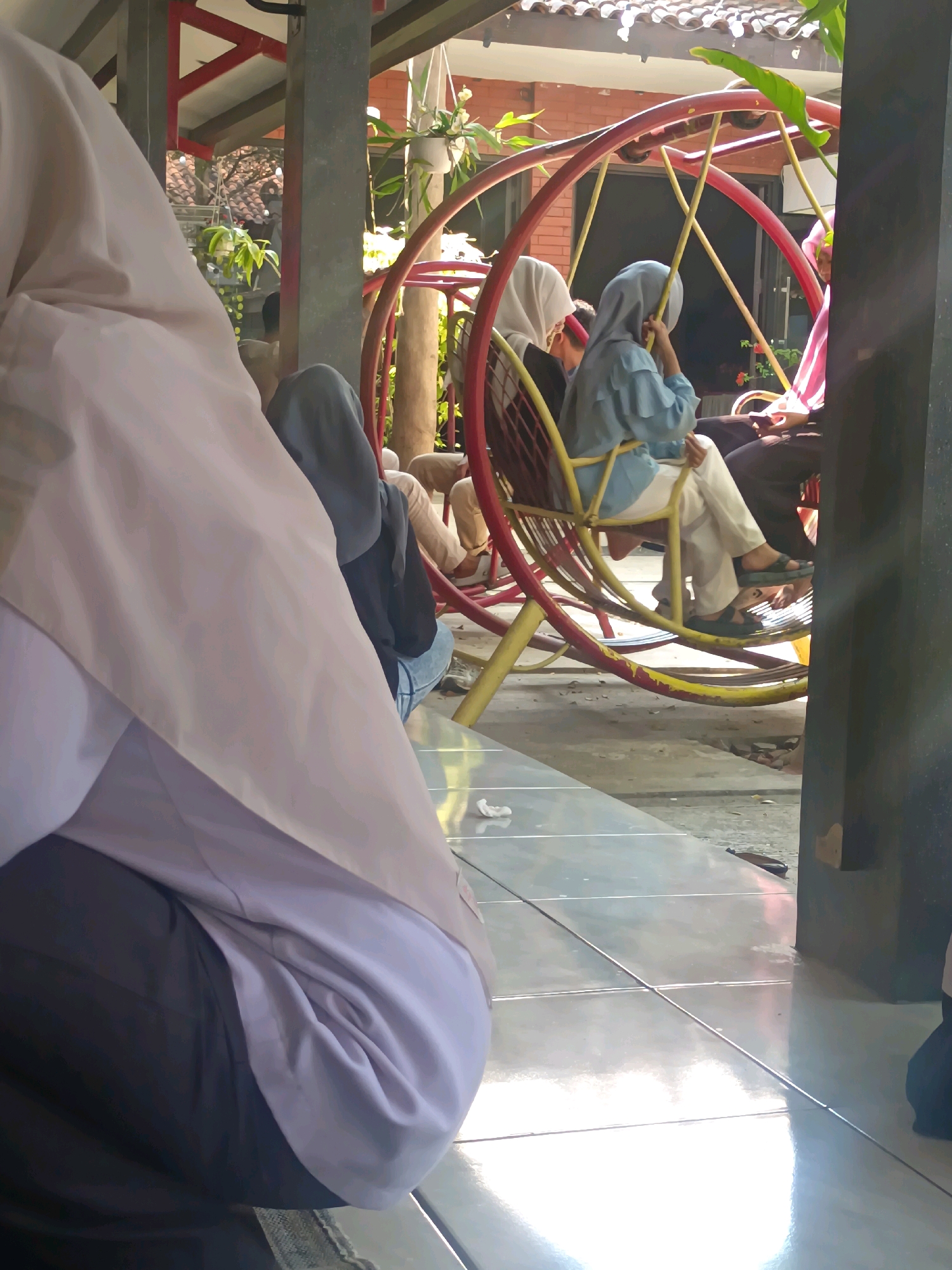 The image depicts a group of people sitting on a circular swing at what appears to be an outdoor leisure area, possibly a park or community space, in Kerangkulon, Indonesia. In the foreground, a person wearing a light-colored hijab and dark pants is partially visible, their back turned towards the camera. Behind them, several other individuals are seated on the swing. One person in the center wears a light blue hijab and white pants, resting their chin on their hand. To their left, another individual in a white hijab is seated. A third person in the swing is partially obscured, wearing a pink and white striped garment and a head covering. The swing itself is red with yellow supports and is positioned on a tiled floor. In the background, potted plants hang, and a building with a tiled roof is visible. The overall scene suggests a casual gathering on a bright day.