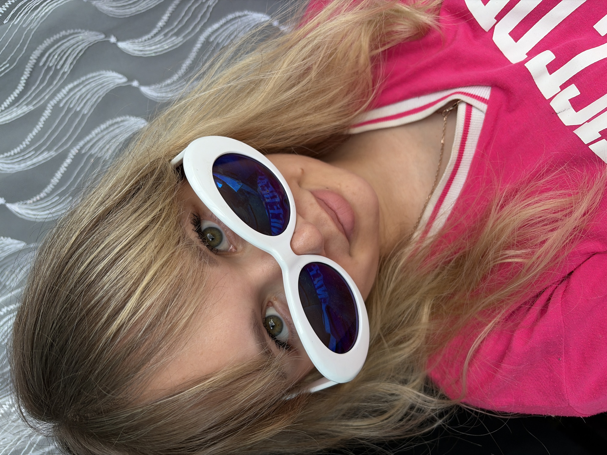 A woman with blonde hair is lying down, wearing white oval sunglasses with blue lenses and a pink sweatshirt with white lettering. The background appears to be a gray textured surface. The location is not identifiable as Chaikovsky, Russia from this image.