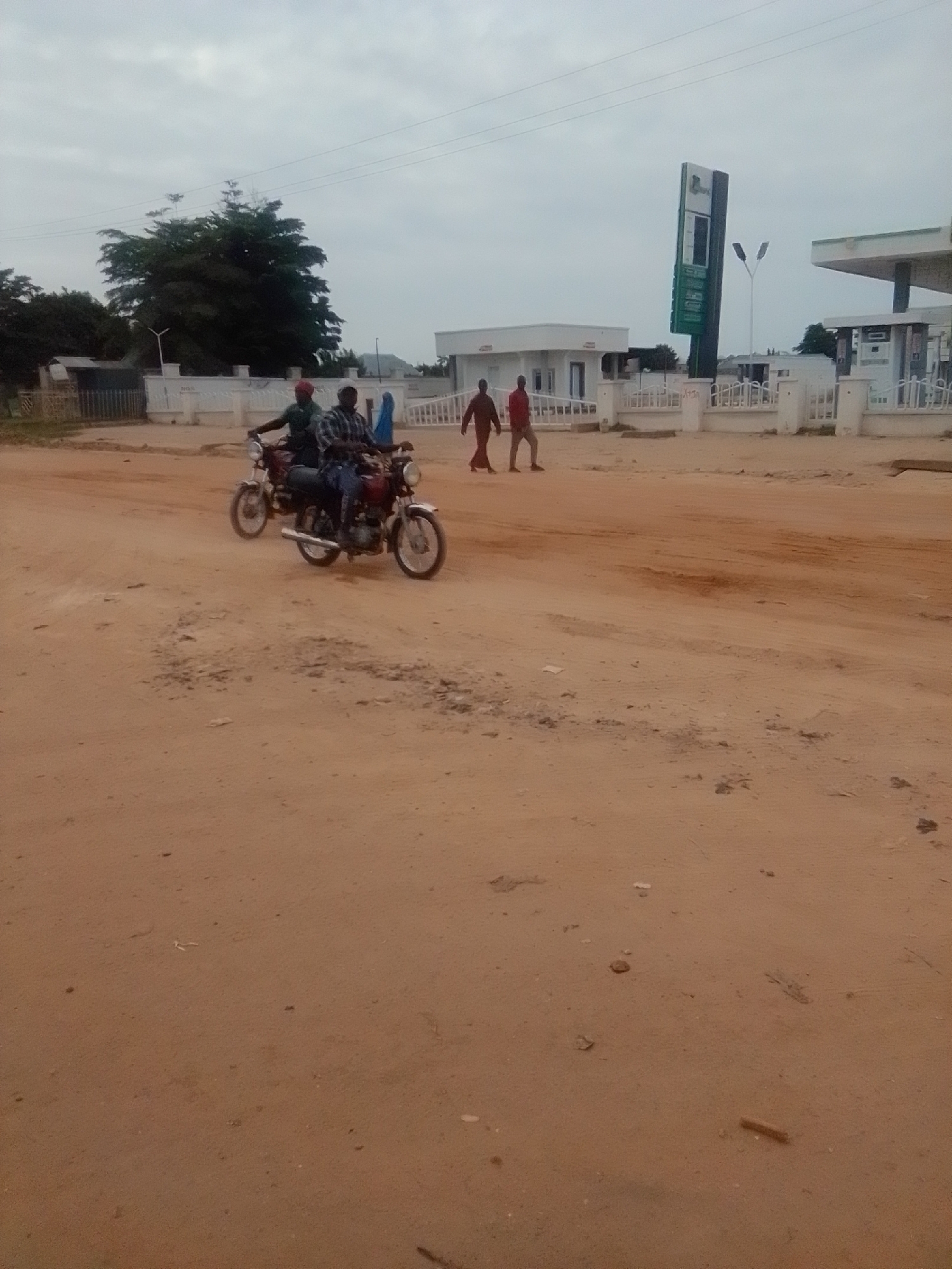 In Gombe, Nigeria, a brown, dusty road features two men riding a red motorcycle. The man in front wears a green shirt and a red cap, while the man behind him is in a plaid shirt. Further down the road, two other men walk, one in a red shirt and the other in a dark shirt. In the background, a white building with awnings stands next to a tall, green sign with white lettering, likely a gas station. The sky is overcast.