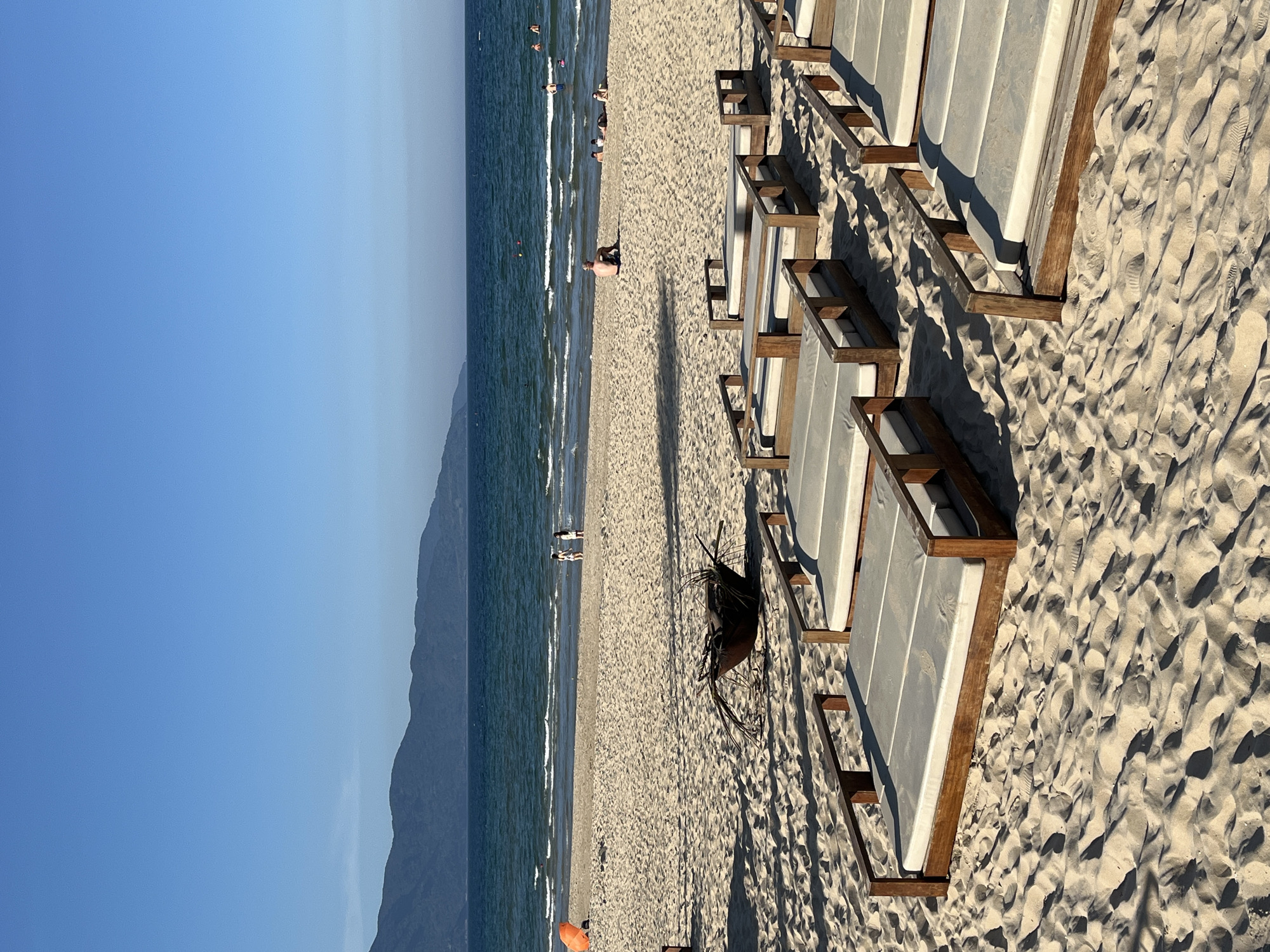 The image shows a beach scene in Đà Nẵng, Vietnam. To the left, there's a clear blue sky above a mountain range. The ocean waves meet the sandy beach, where a few people are swimming and sunbathing. On the right side of the image, there are rows of empty beach lounge chairs with white cushions and wooden frames placed directly on the sand. There is also a dark plant lying on the sand between the lounge chairs and the beach.