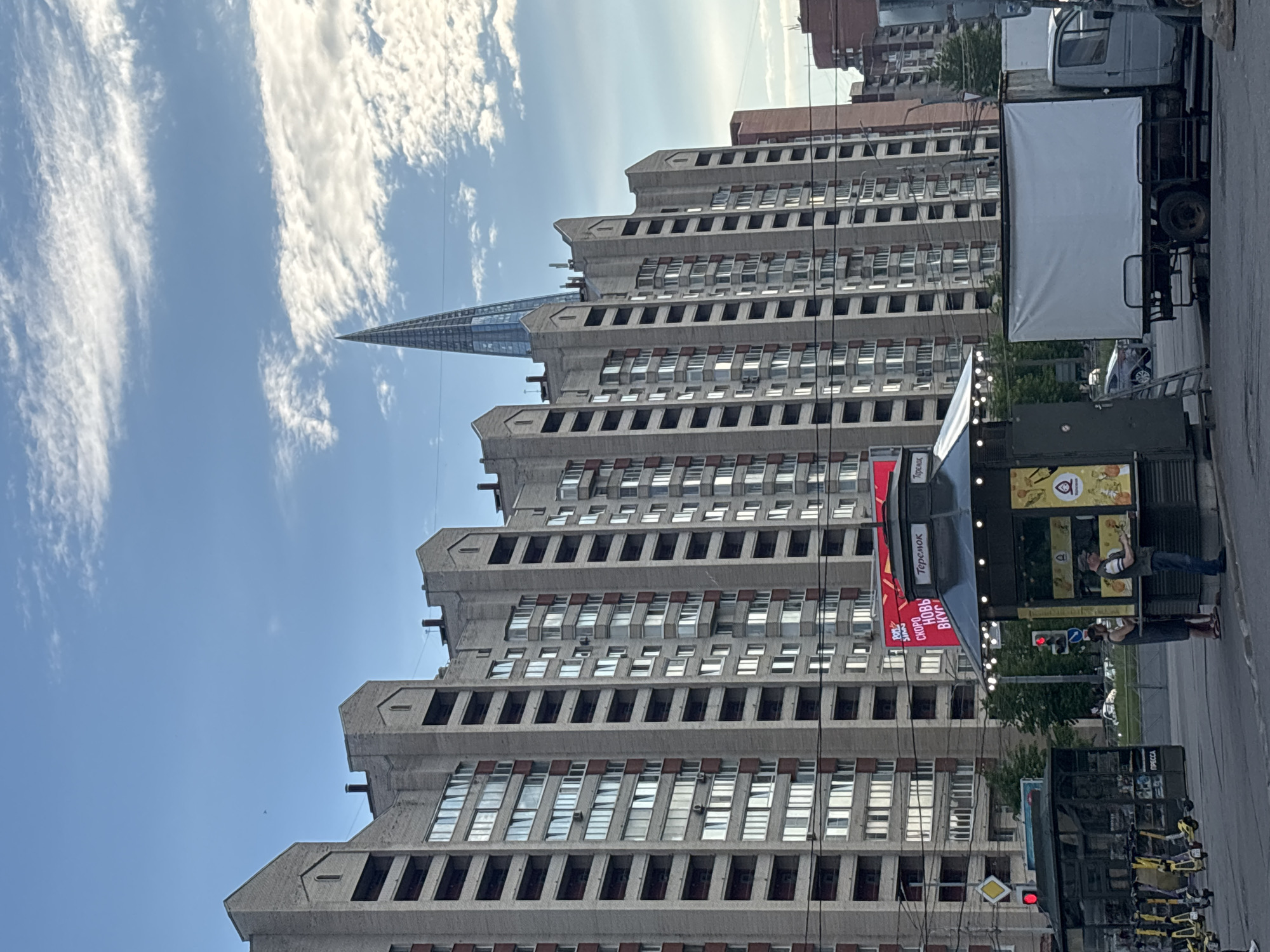 This outdoor, vertically-oriented image captures a busy urban street scene in St. Petersburg, Russia, likely during the daytime under a partly cloudy sky. The most prominent subject is a tall, brutalist-style multi-story building, possibly residential or office, with numerous rectangular windows and distinctive, tiered concrete elements forming its upper levels. Its weathered concrete facade suggests age.
In the background, beyond the brutalist building, the sleek, glass-clad spire of the Lakhta Center skyscraper is clearly visible, rising sharply against the blue sky and scattered white clouds. This landmark confirms the location as St. Petersburg.
At street level, the scene is bustling with everyday activity. On the right, a large truck with a white canvas cover is parked. Adjacent to the building, several kiosks or stands are present. One black-colored kiosk features a red banner with the text "Теремок" (Teremok, a Russian fast-food chain), "Мы скоро с НОВЫЙ ВКУС" (We are soon with NEW TASTE). Another kiosk has yellowish panels and visible text "ПИВО" (Beer), "ХМЕЛЬНОЙ КОЛОС" (Hoppy Spikelet), "Жареные креветки" (Fried Shrimp), and "Торговый ряд" (Shopping row).
Several people are visible near these kiosks, including one person standing by the yellow-paneled kiosk and another further back. A row of yellow rental bicycles or scooters is parked on the sidewalk. A red traffic light and a yellow diamond-shaped street sign are also present. The overall impression is one of a functional, lived-in city environment.