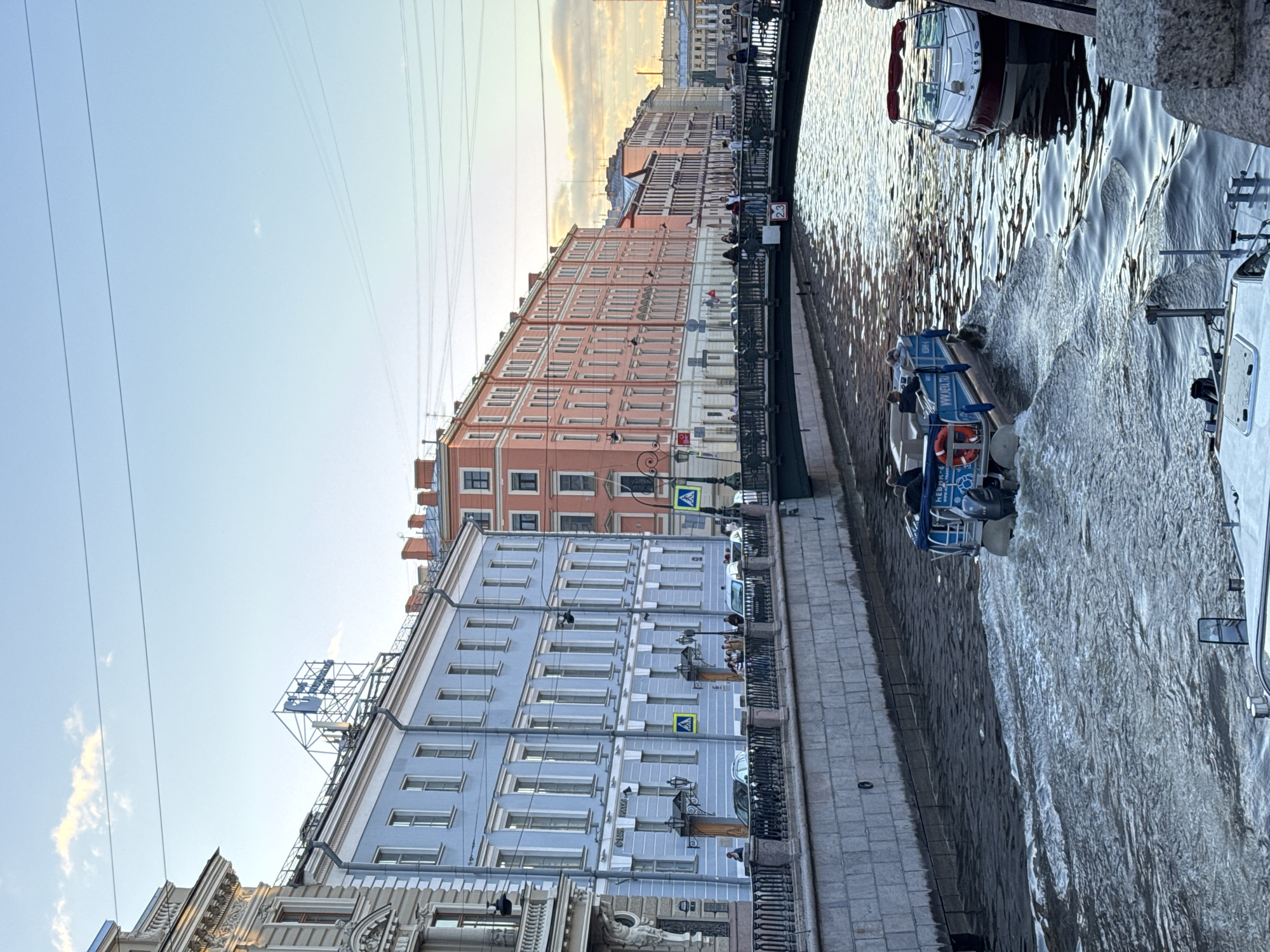 This image depicts a picturesque canal scene in the city of Imeni Morozova, Russia. The canal is lined with elegant buildings, featuring a light blue facade on the left and a series of salmon-pink buildings extending to the right, all showcasing traditional European architecture with numerous windows and decorative elements. A pedestrian bridge spans the canal, with people visible walking across it.
On the water, a blue tour boat with several passengers is actively moving, creating ripples and wakes. Another white boat with a red canopy is docked on the right side of the canal. The canal walls are constructed of stone blocks, and a metal railing runs along the embankment. The sky above is a clear blue with a few wispy clouds, illuminated by the soft light of what appears to be late afternoon or early evening sun. Overhead, power lines are visible, a common feature in urban landscapes. The overall impression is one of a tranquil urban setting, showcasing the city's architectural charm and canal system.