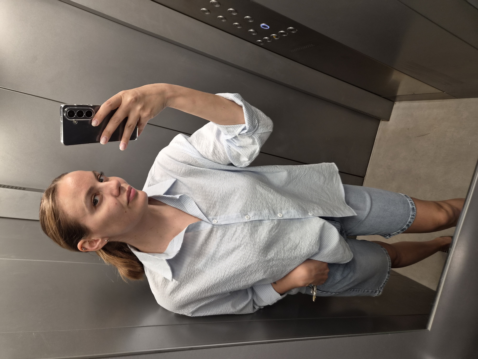 A woman takes a selfie in an elevator. She holds a black phone in her right hand, which has three camera lenses visible. The woman has blonde hair pulled back in a ponytail and is wearing a light blue and white striped button-down shirt with denim shorts. She's standing in the elevator, and the elevator button panel is visible above her. The interior of the elevator is a gray, metallic finish. There's no evidence to link this picture to Chaikovsky, Russia.