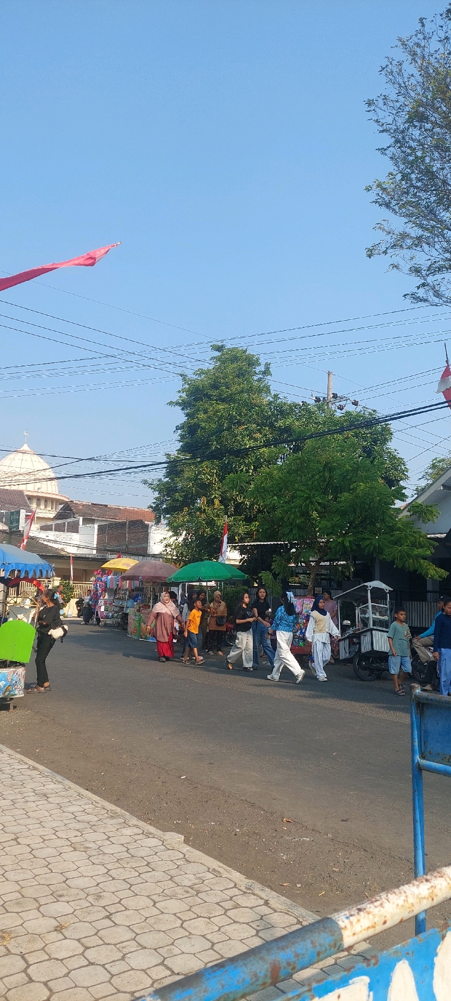 The image depicts a street scene in Jogoyudan, Indonesia, on a clear day with a blue sky. The street is lined with several vendor stalls covered by colorful umbrellas, offering various goods. A diverse group of people, including women in hijabs and children, are walking along the street, browsing the stalls, and interacting with each other. Indonesian flags are visible, adding a patriotic element to the scene. In the background, a building with a dome, possibly a mosque or a similar structure, can be seen. The presence of power lines overhead indicates an urban or semi-urban setting. The foreground shows a paved sidewalk and a section of a metal barrier.