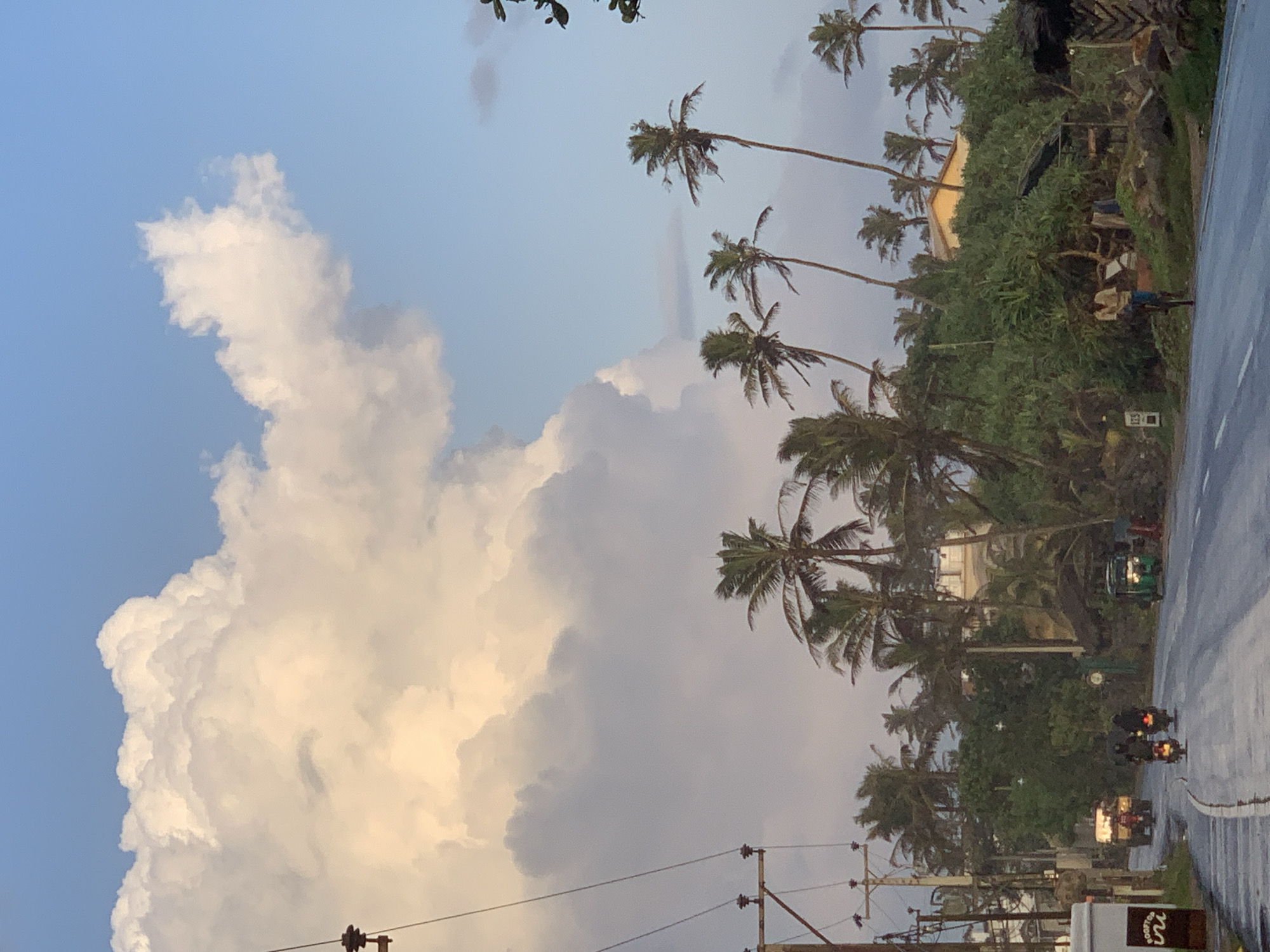 In Ahangama, Sri Lanka, a paved road runs through a tropical landscape under a sky dominated by dramatic clouds. The clouds are large and billowy, with a bright yellow hue on the left side, suggesting either sunrise or sunset. The sky transitions to a clear, pale blue on the right. Palm trees are abundant throughout the scene, their fronds swaying gently.
On the road, several motorbikes are visible, with riders moving in the same direction. A three-wheeled auto-rickshaw is also present, its lights illuminated. A truck is further up the road. A lone man rides a bicycle on the side of the road, and another person stands near the rickshaw.
Along the roadside, there are signs, electrical poles with wires, and dense green foliage. Buildings, including a white structure on a hill and what appears to be a thatched-roof hut, are nestled among the vegetation. The road surface is wet, indicating recent rain. A sign with the number "125" is visible on a post.