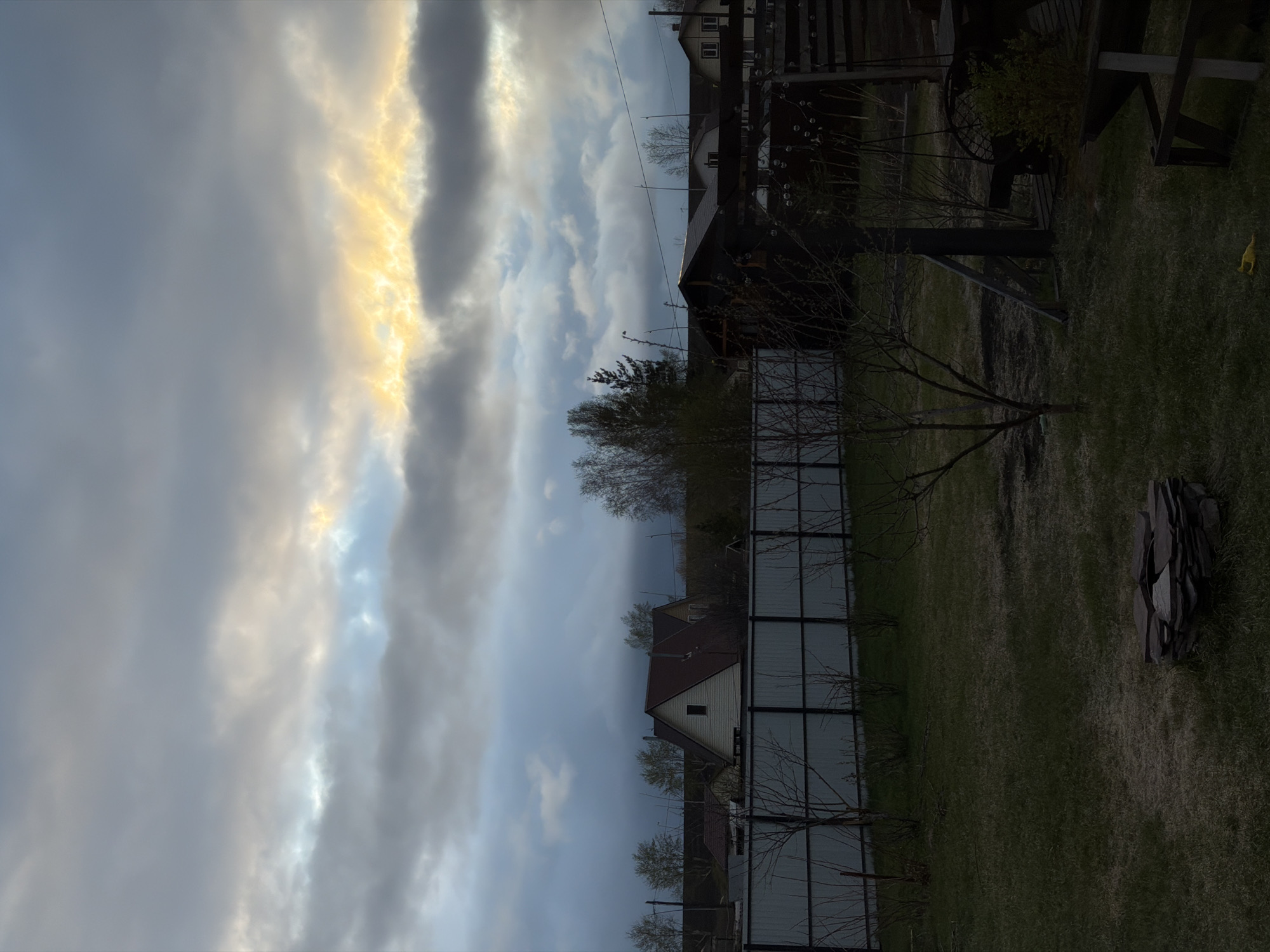 The image shows a residential backyard in an indeterminate city location. The yard features a metal fence, a patch of green grass, and several small trees with bare branches. In the distance, houses with dark roofs are visible. The sky is filled with gray clouds, and a band of golden light is breaking through. There are no people in the image. The environment is suggestive of a late afternoon or early evening.