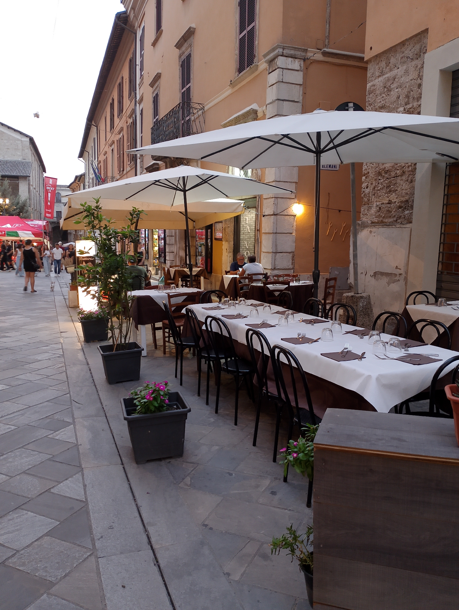 The image depicts an outdoor seating area of a restaurant on a cobblestone street. Several tables are set with white tablecloths, glasses, and cutlery, some covered by large white umbrellas. Plants in dark pots are placed around the tables. In the background, people are walking along the street, and buildings with shuttered windows line the area. The street appears to be part of the historic center of Rieti, Italy, with traditional architecture and a pedestrian-friendly atmosphere. The lighting suggests it is late afternoon or early evening.