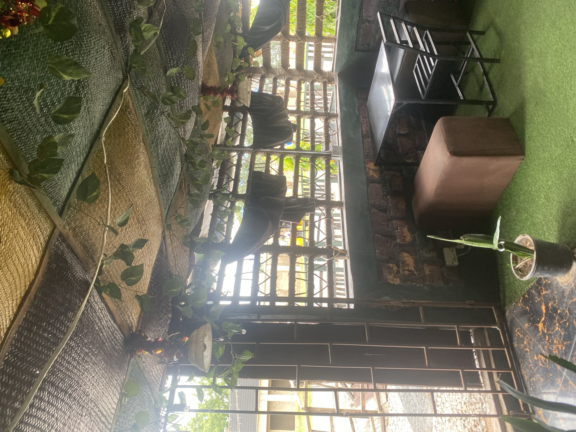 This image captures an outdoor seating area, likely a cafe or restaurant, with a rustic and natural aesthetic. The scene is set during the day, with sunlight filtering through a lattice-like structure, creating patterns of light and shadow. The overall feel is relaxed and somewhat bohemian.
The primary subjects are the various elements of the seating area. Woven mats in shades of brown and green form a textured backdrop, draped and layered to create an enclosure or wall. Lush green artificial vines with broad leaves are woven through this material, adding a touch of nature. In the background, a metal grate or trellis with a rough, rope-like texture runs horizontally, with more sunlight and foliage visible beyond it.
Dominating the right side of the image is a simple black metal table with a reflective top, and next to it, a plush brown ottoman. The ground is covered in artificial green turf, typical of an outdoor or semi-outdoor recreational space. A small potted plant with slender green leaves sits on the turf near the table. In the distance, through the grate, glimpses of buildings and trees suggest an urban or semi-urban environment. The lighting suggests it is daytime, and there are no obvious weather cues.
There is no visible text in the image. The scene evokes a sense of a tranquil, green retreat, perhaps a place to relax or socialize in a comfortable, nature-inspired setting.